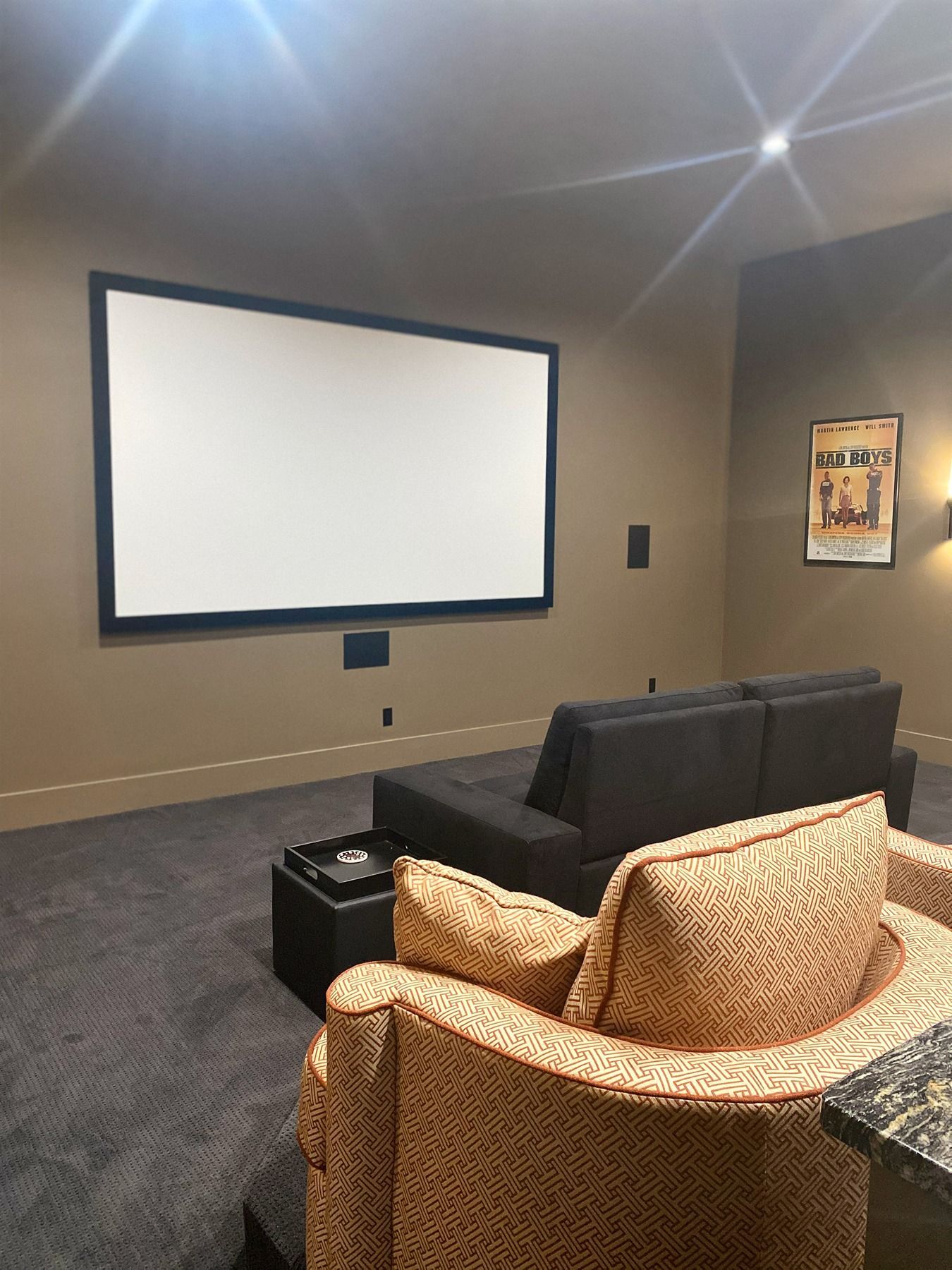 A home theater featuring a large screen on a brown wall, a dark gray sofa, and a patterned beige armchair.