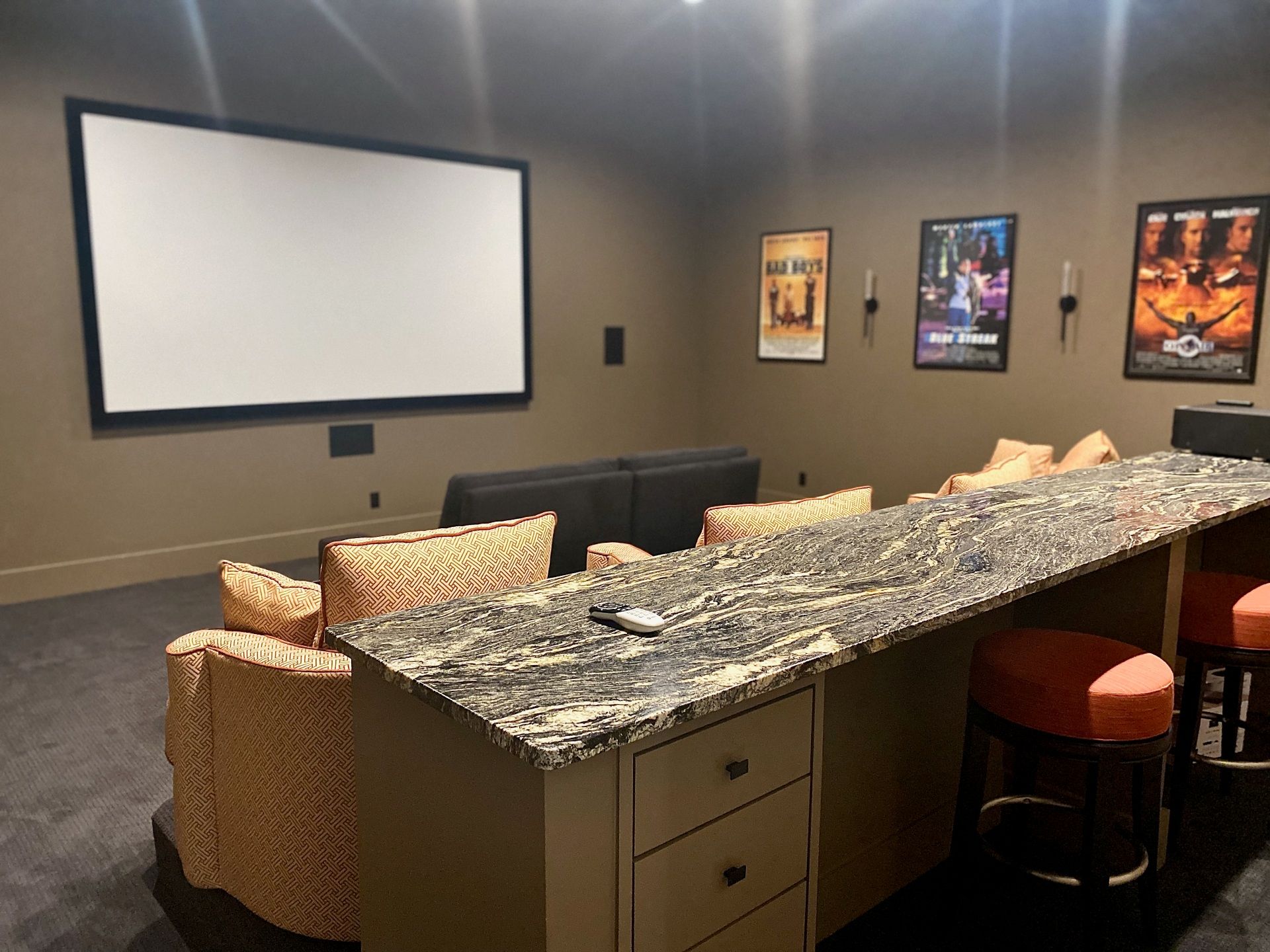 A home theater with a large screen, tan sofas, and a granite-topped bar with orange stools against a dark wall.