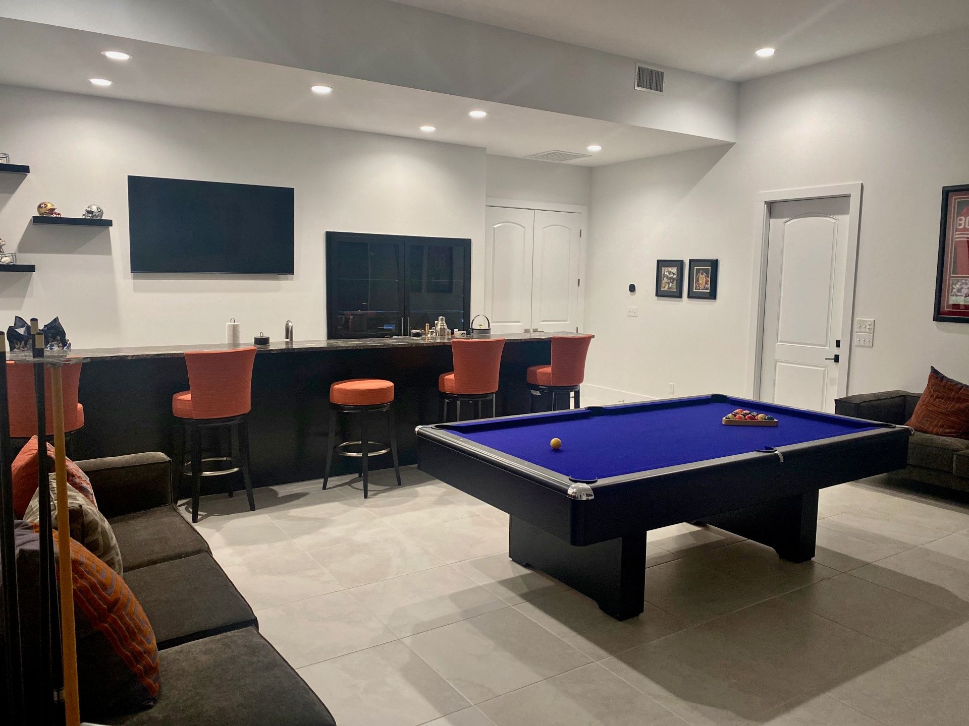 A basement lounge with a blue pool table, a bar with three orange stools, and a seating area with a sofa.