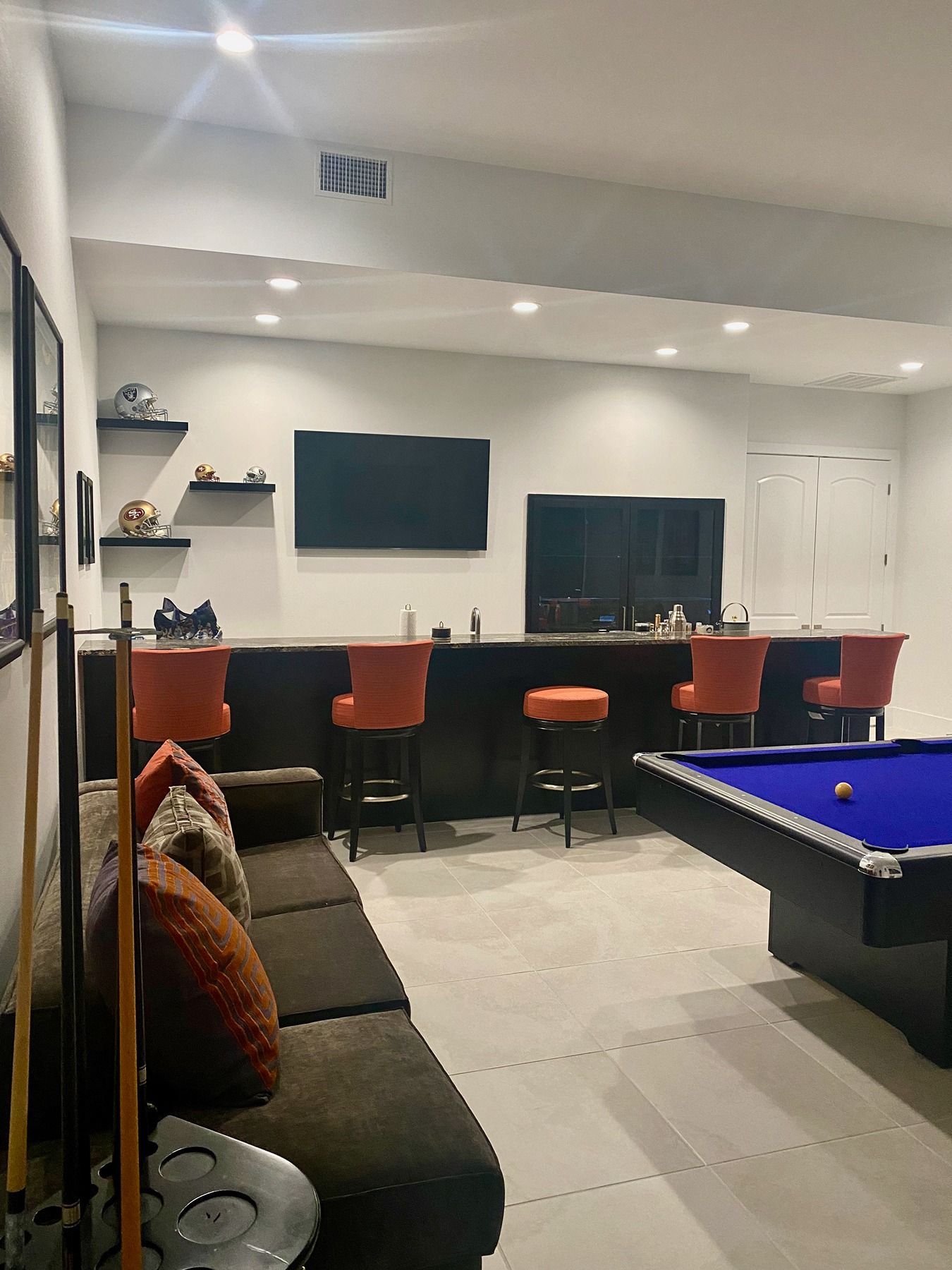 A room featuring a pool table in the foreground, a bar with orange stools, a TV, and shelves on the back wall.