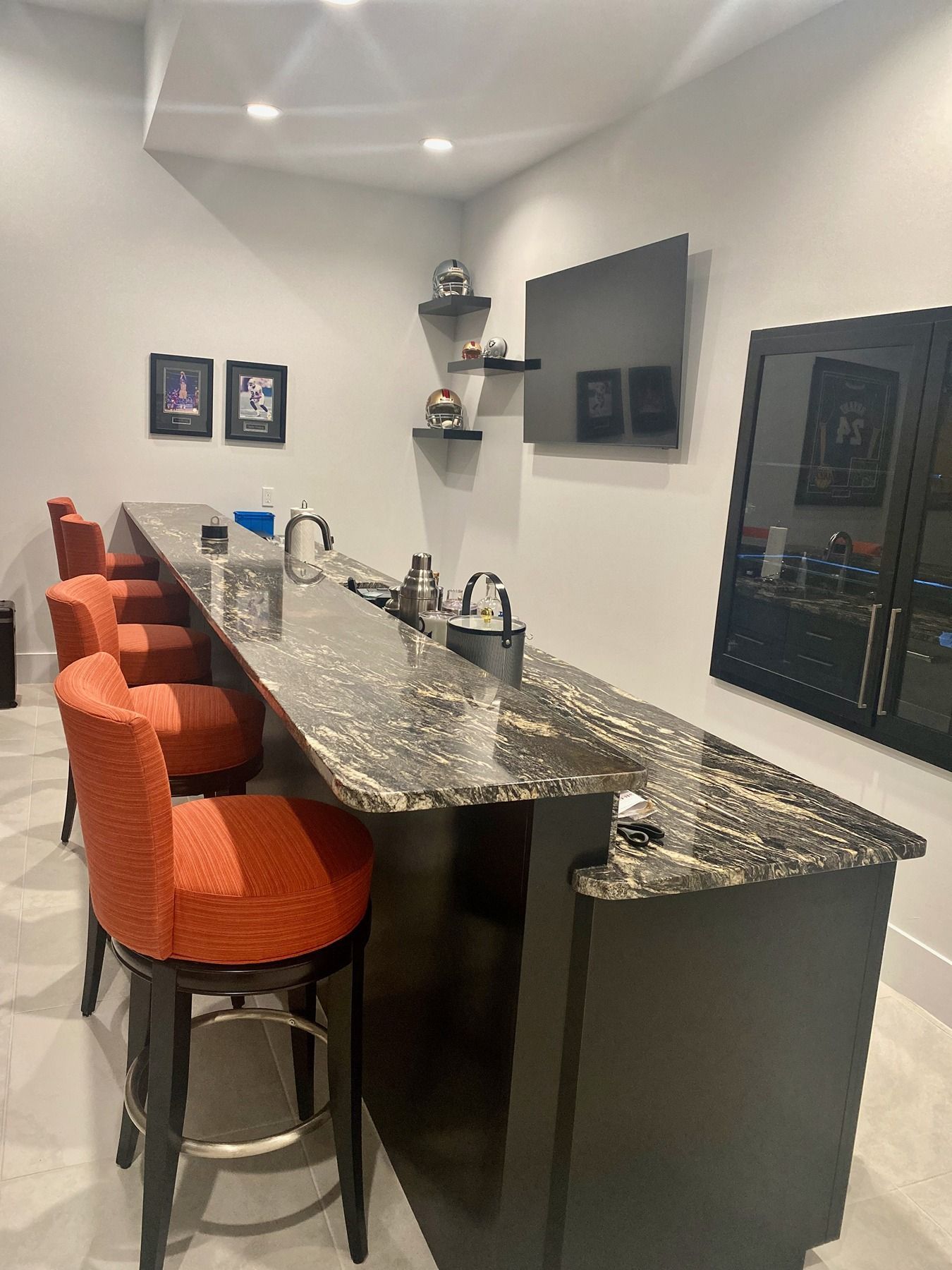 A sleek home bar featuring dark wood cabinets, a marble countertop, orange bar stools, and a wall-mounted TV.