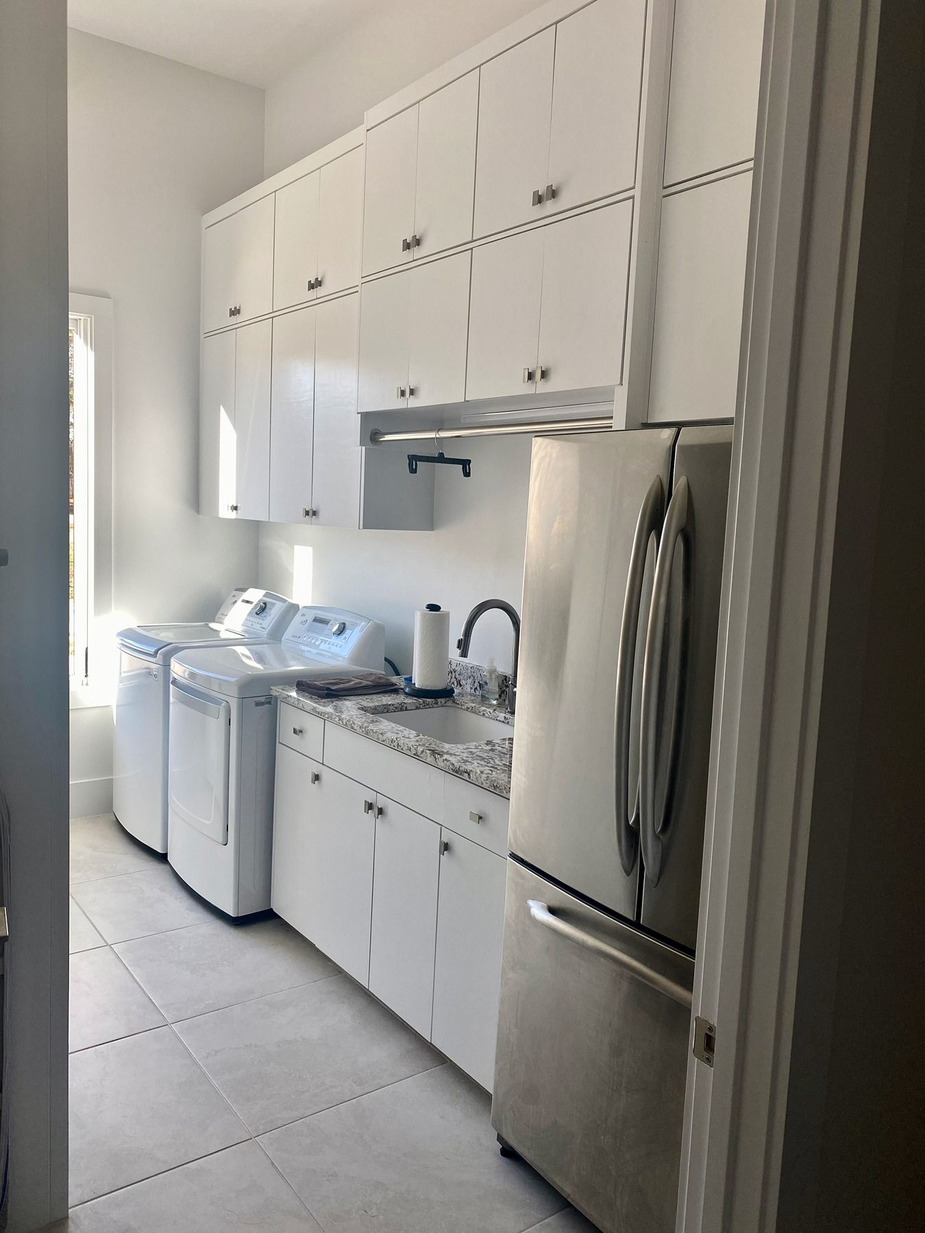 A laundry room with white cabinets, a sink, granite countertops, a stainless steel refrigerator, and two white appliances.