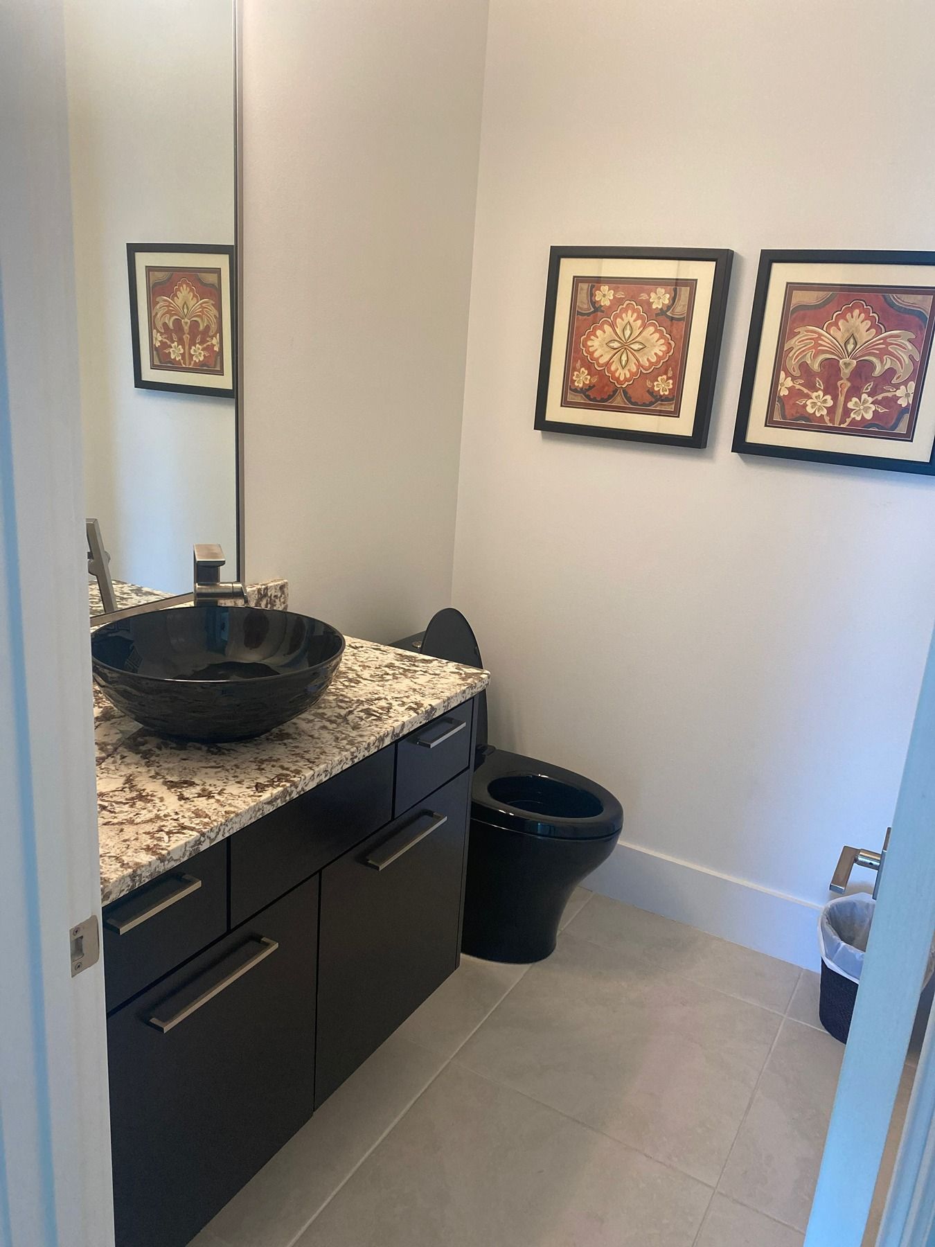 Modern bathroom with a black vessel sink on a granite counter, dark cabinets, a black toilet, and two framed wall prints.