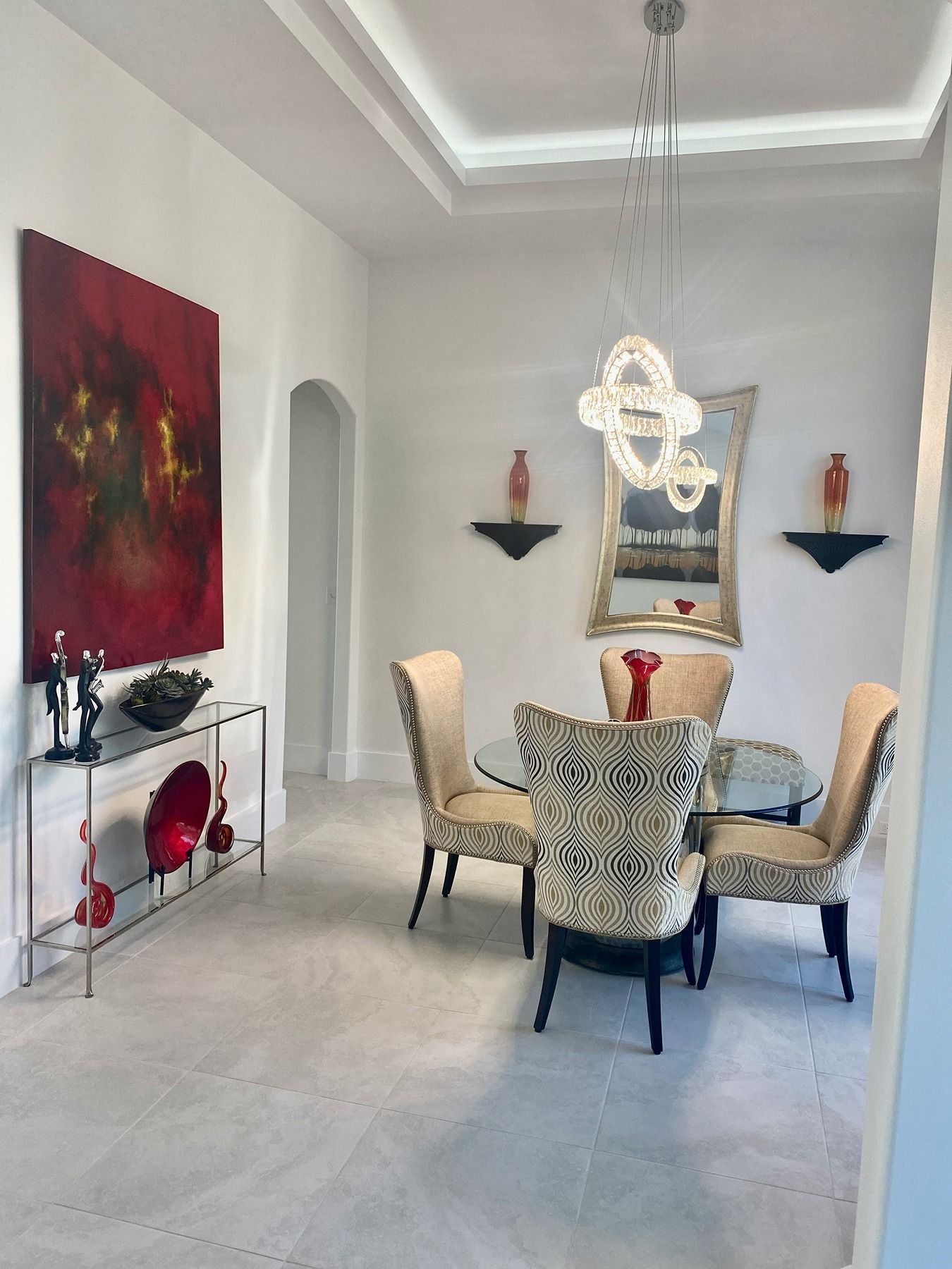 A dining room with a glass table, four patterned chairs, a red abstract painting, and a crystal chandelier.