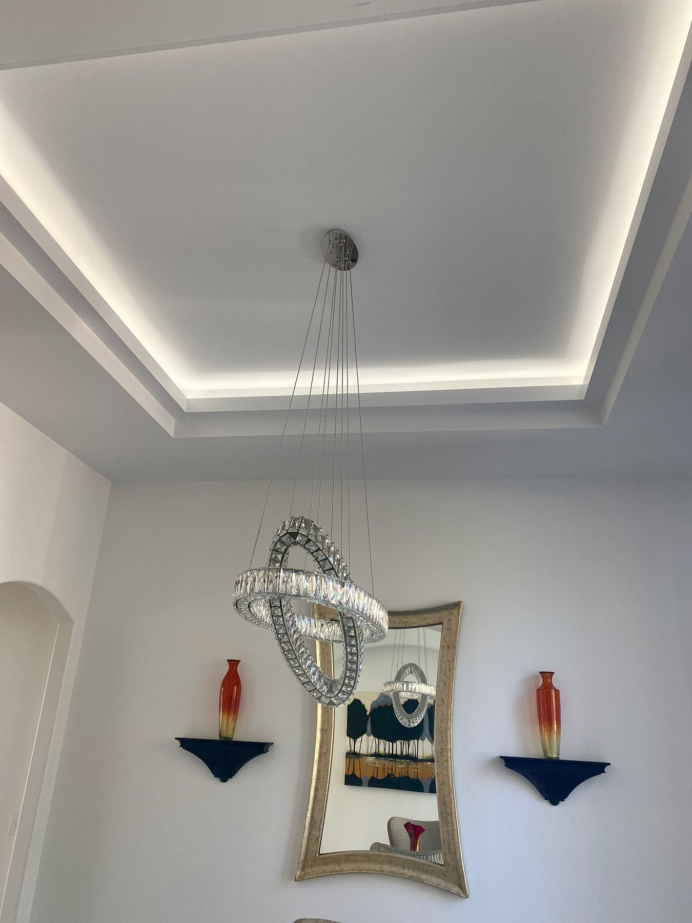 A modern crystal ring chandelier hangs from a recessed tray ceiling with LED cove lighting above a gold-framed mirror.