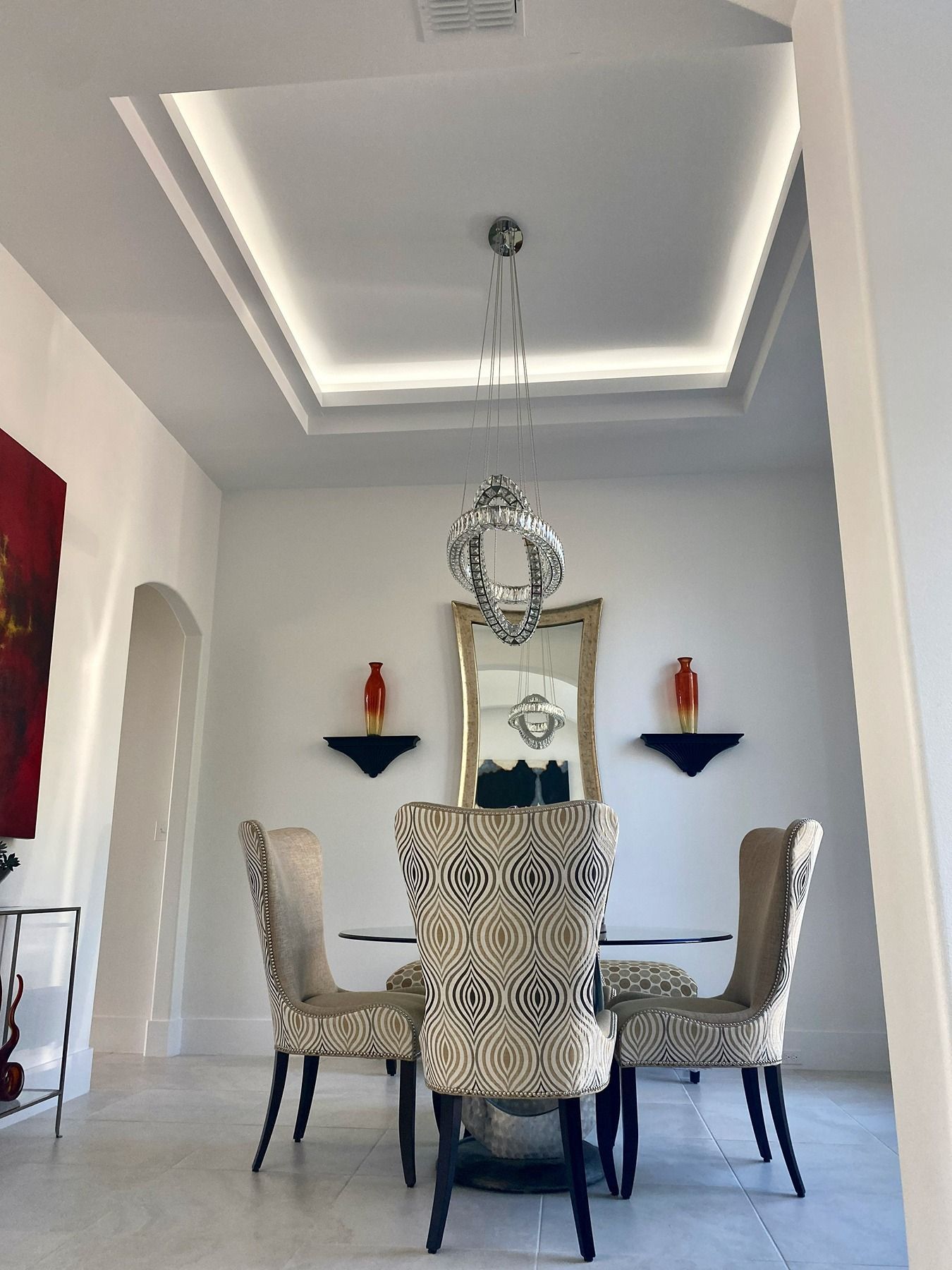A formal dining room featuring a glass table, patterned chairs, a lighted tray ceiling, and a mirror with two wall sconces.