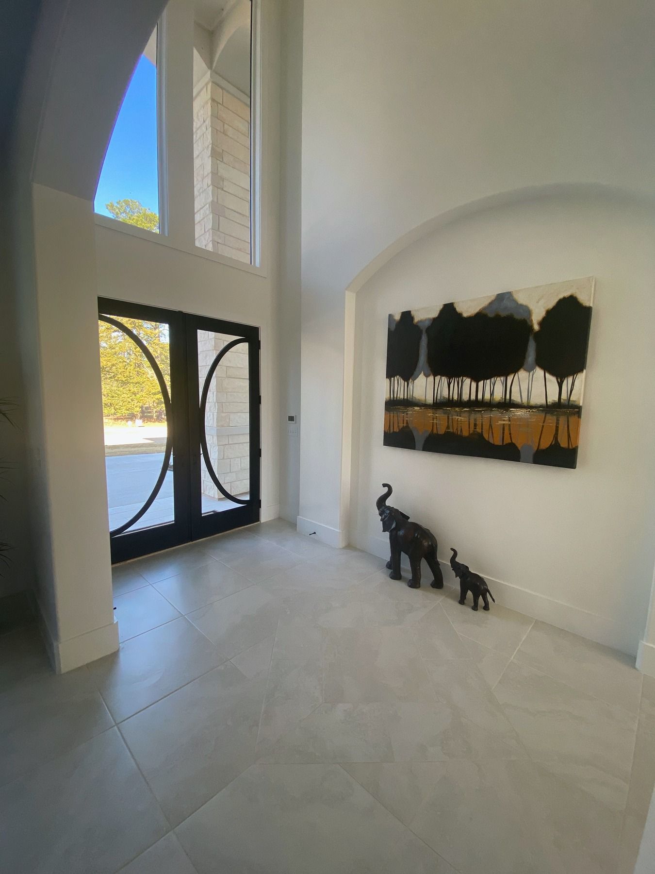 A bright entryway with large glass doors, tile flooring, an abstract landscape painting, and two decorative elephants.