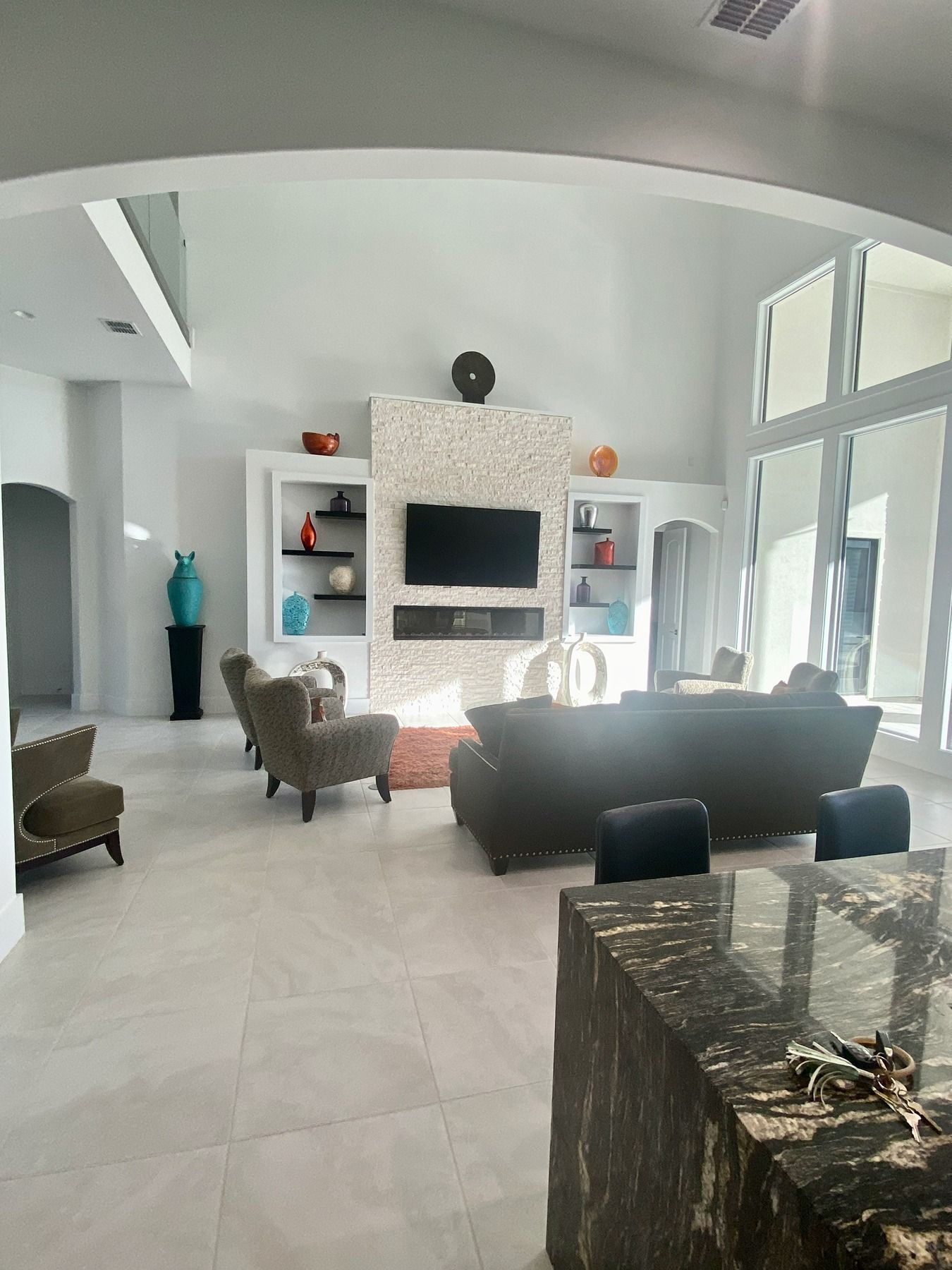 A bright living room with tall ceilings, white walls, large windows, a stone fireplace, and dark-colored furniture.