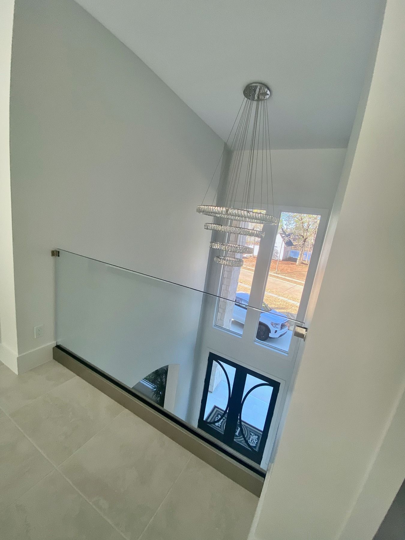 A modern glass railing overlooks a bright foyer with a chandelier and a view of an outdoor street through a tall window.