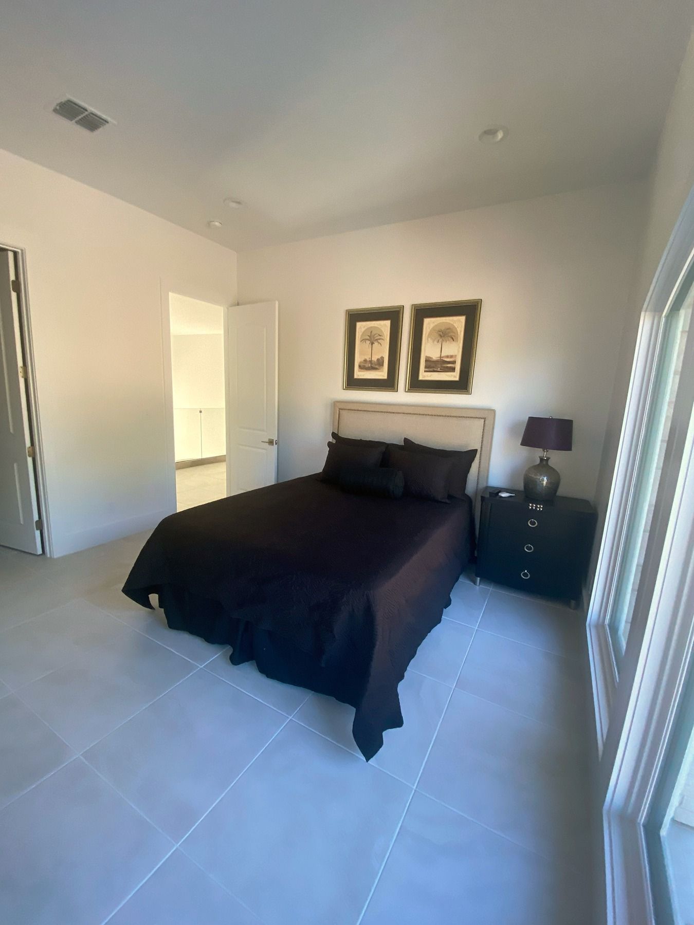 A modern bedroom with a black bed, a bedside table with a lamp, two framed pictures, and tile flooring.