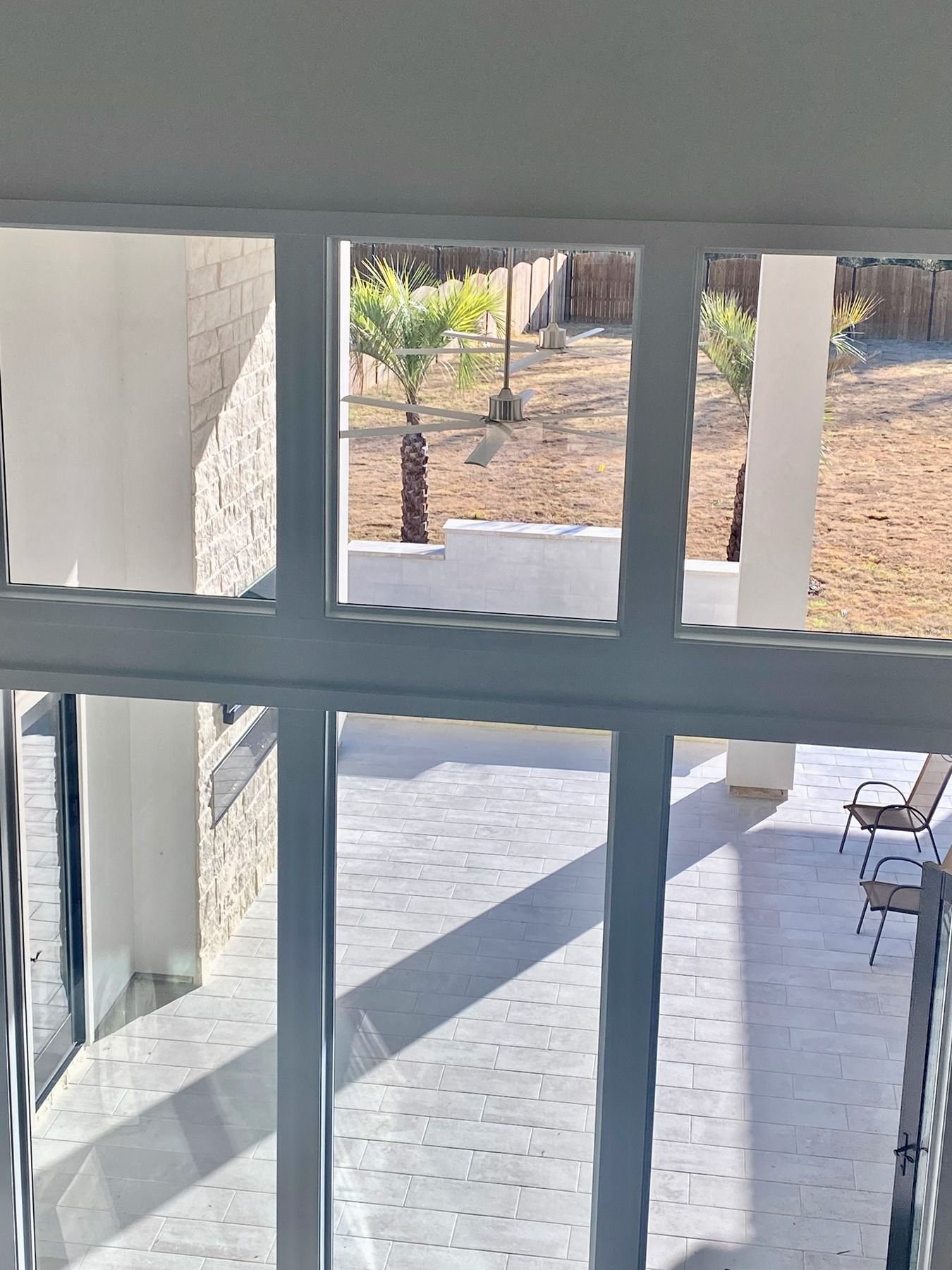 A view through a white-framed window looking out onto a tiled patio with chairs and a sunny yard with small palm trees.