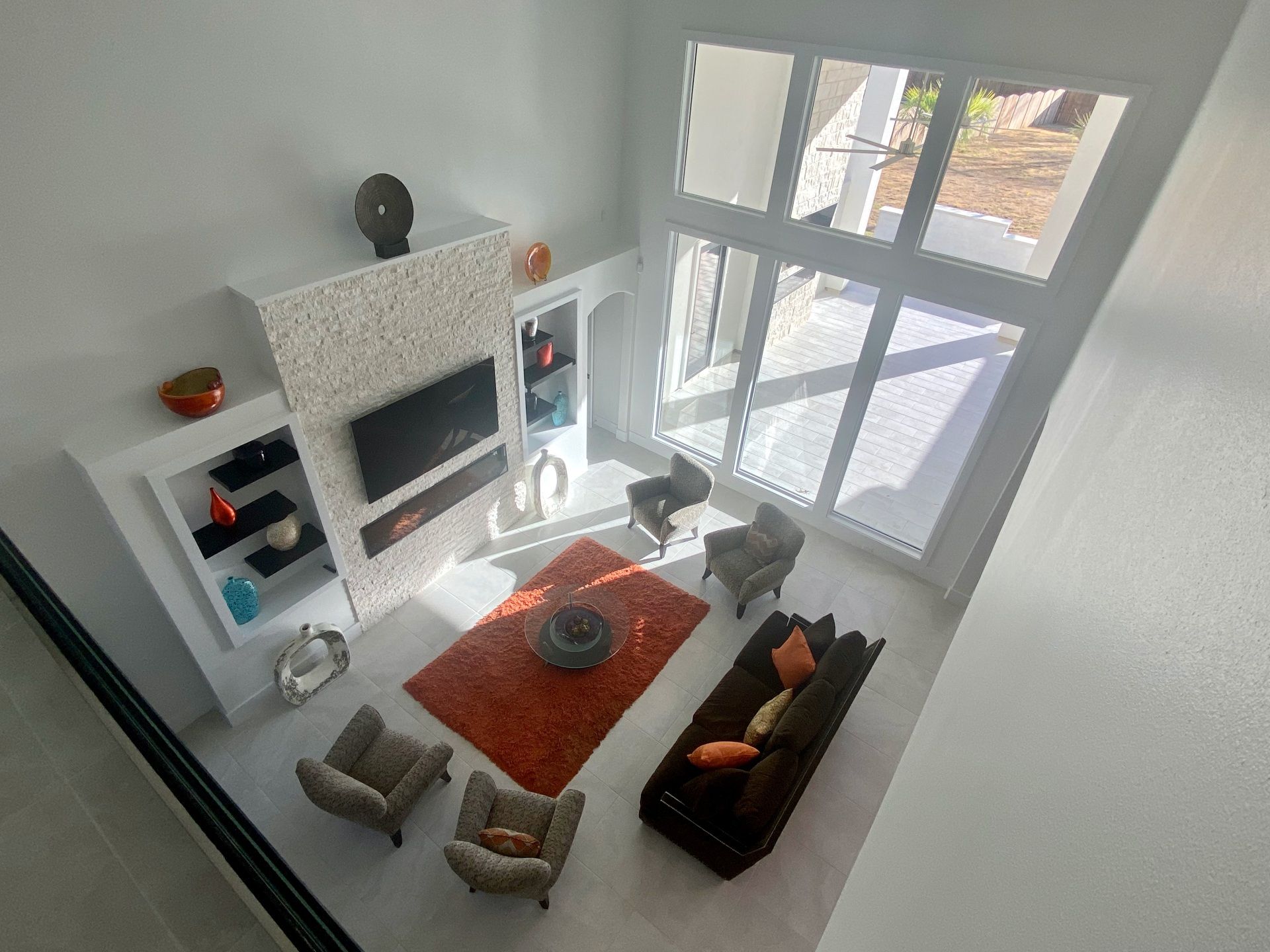 High-angle view of a modern living room with a stone fireplace, orange rug, gray armchairs, and a large window wall.