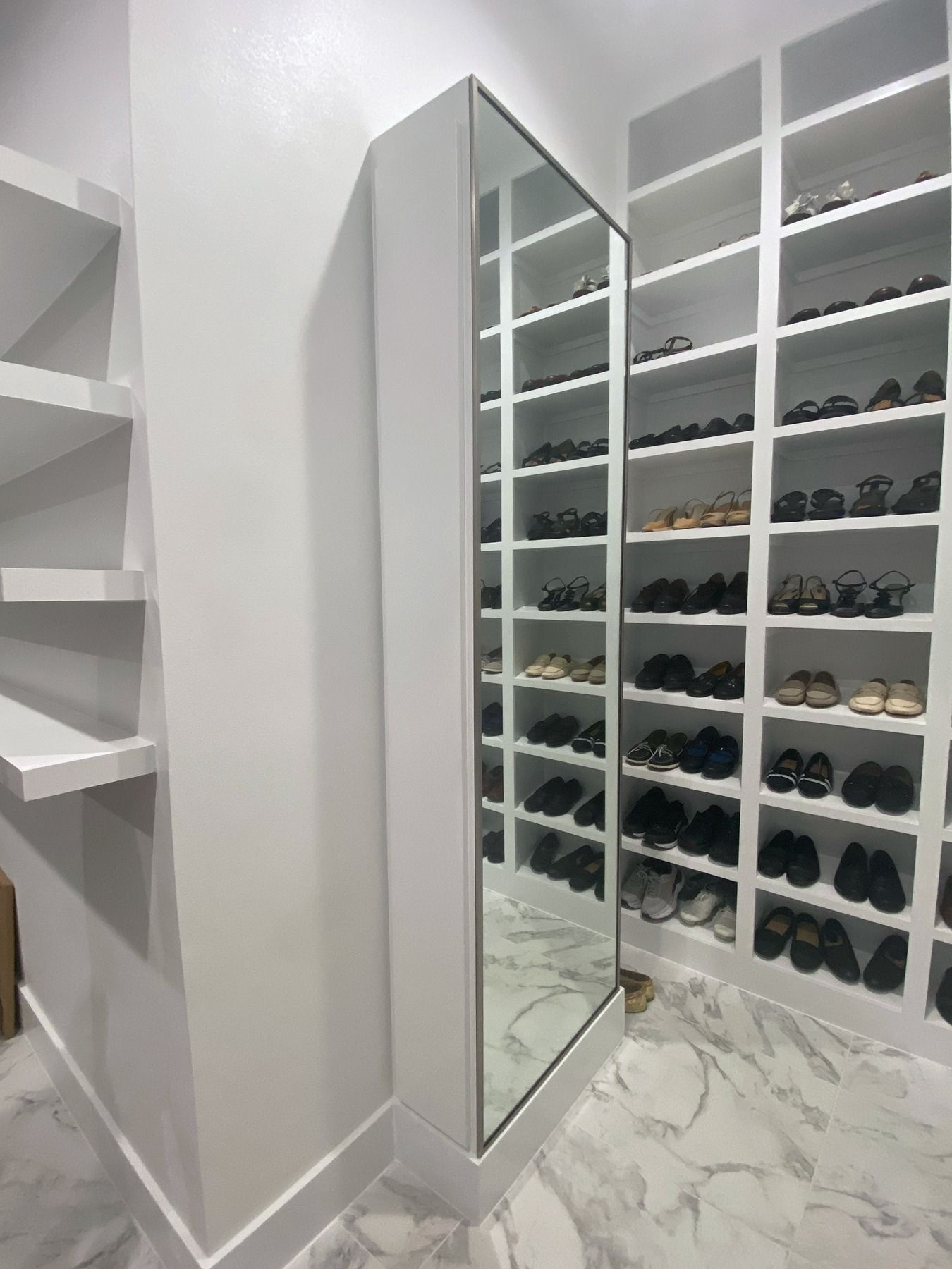 A modern closet with white shelving units, a mirrored cabinet, and marble-patterned flooring filled with various shoes.