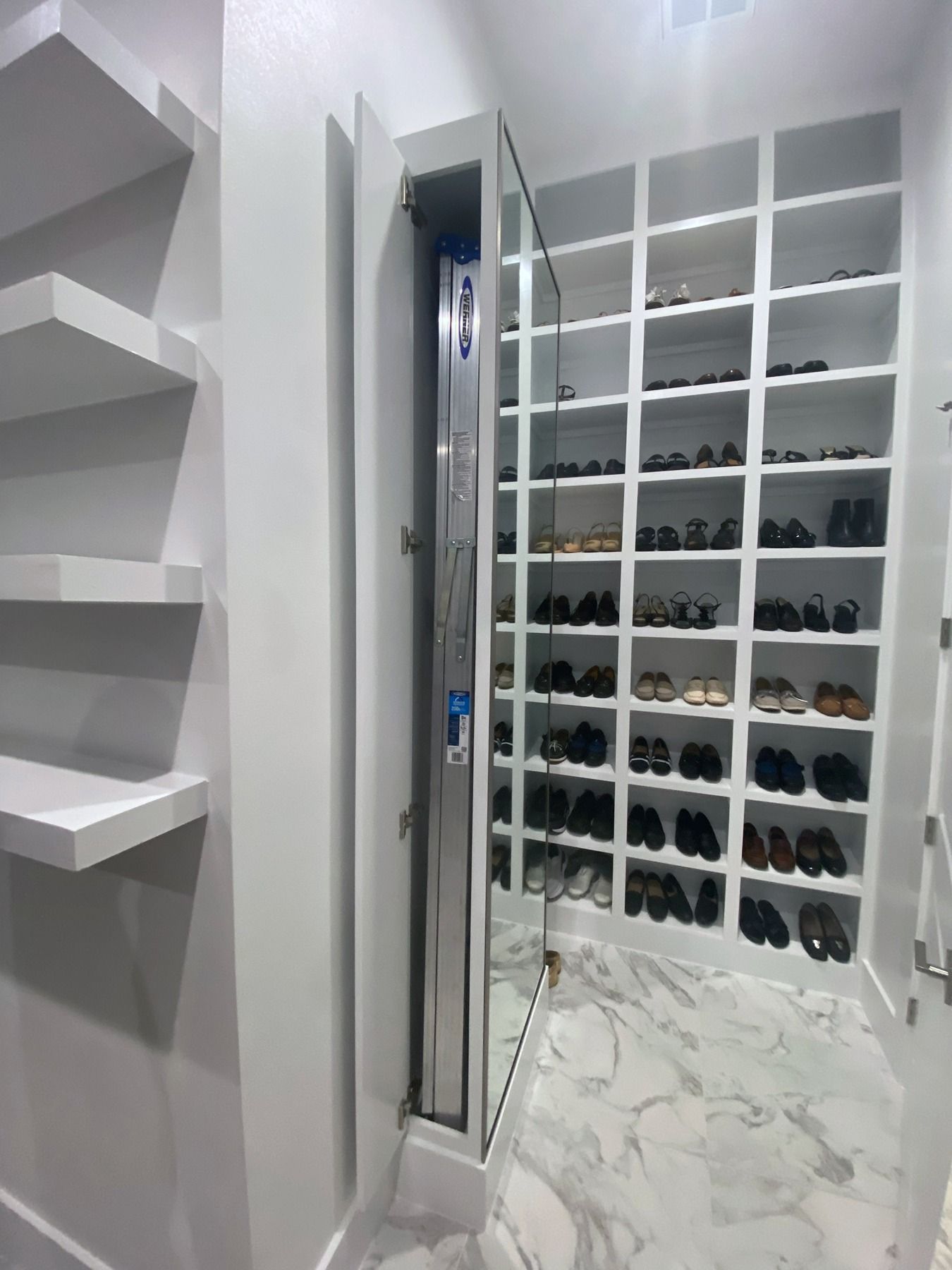 A white walk-in closet with marble floors, featuring tiered shoe shelves and a hidden, vertical ladder storage compartment.
