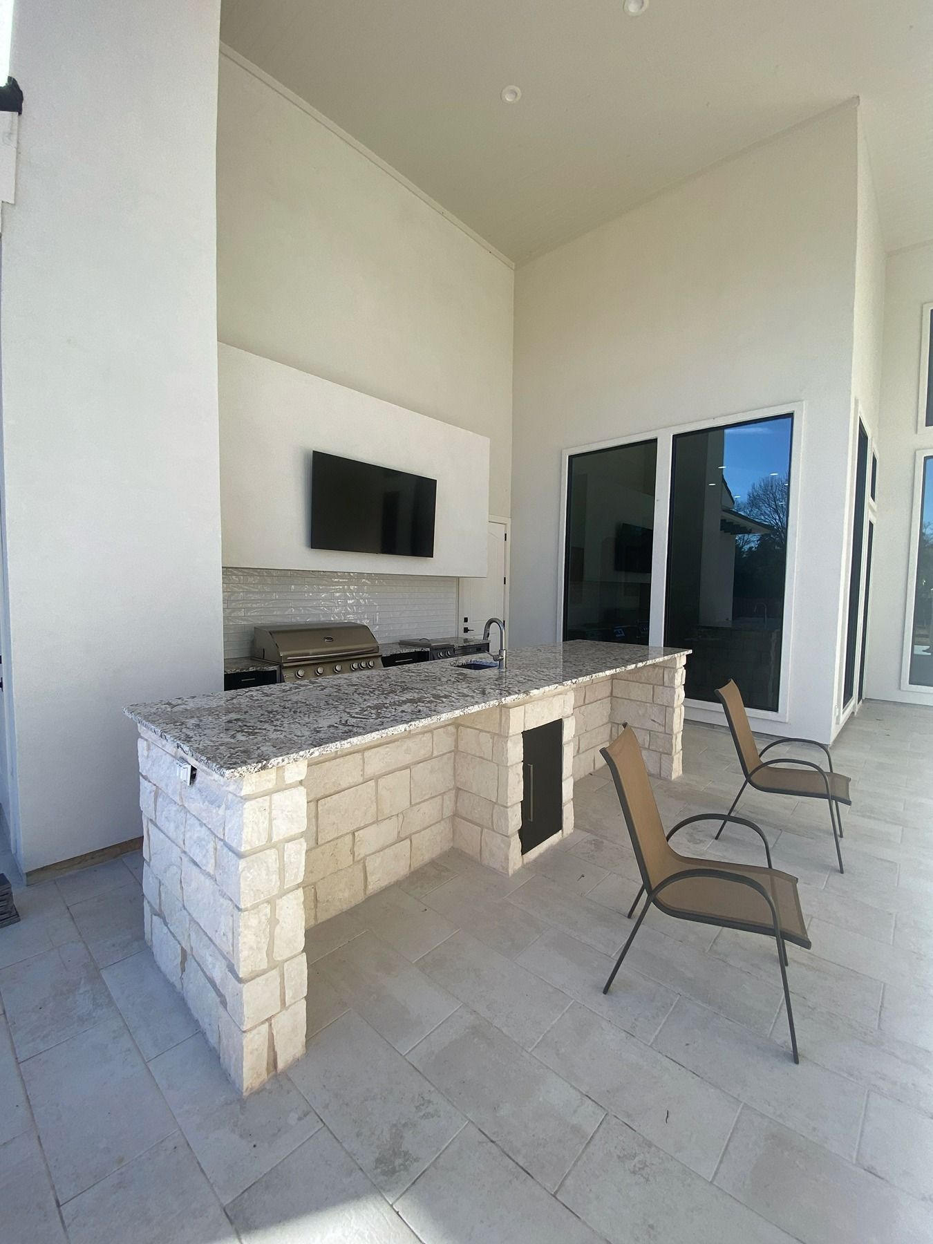 An outdoor kitchen island with a granite countertop and light stone base, featuring a built-in grill and TV, on a patio.