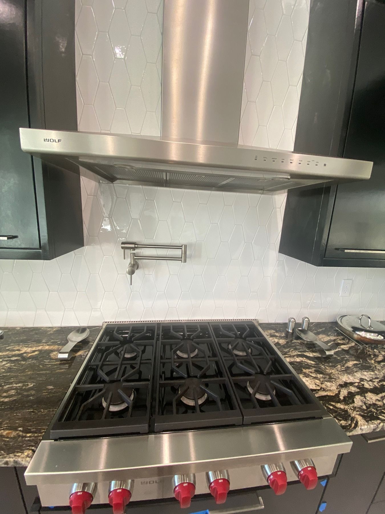 A stainless steel Wolf gas range and vent hood in a kitchen with dark cabinets and a dark stone countertop.