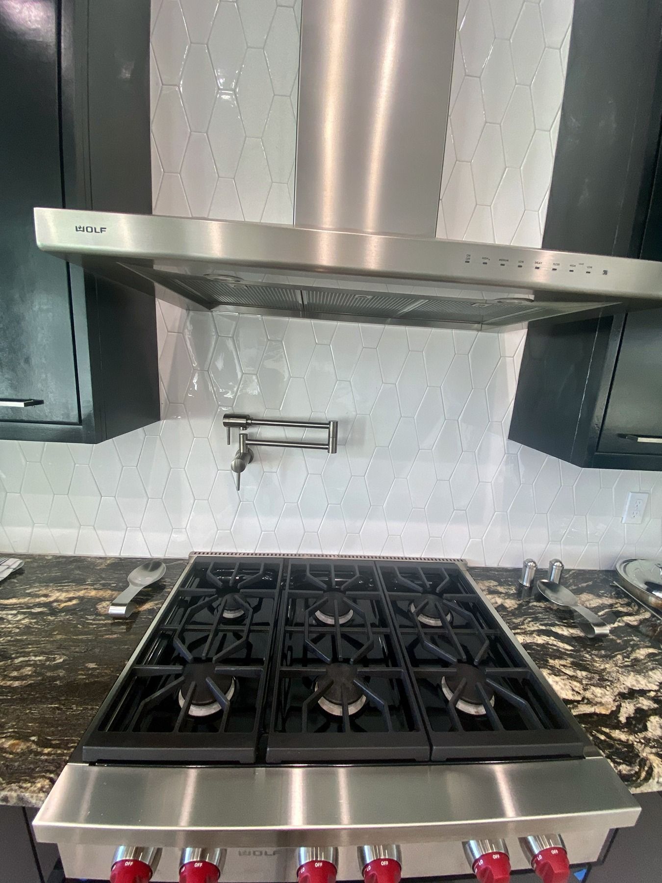 A stainless steel professional gas stove and range hood in a kitchen with black cabinets and a patterned tile backsplash.