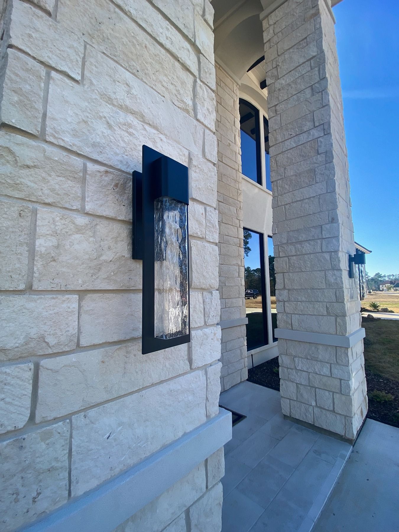 A modern black wall sconce with textured glass mounted on a light-colored stone pillar at the entrance of a house.