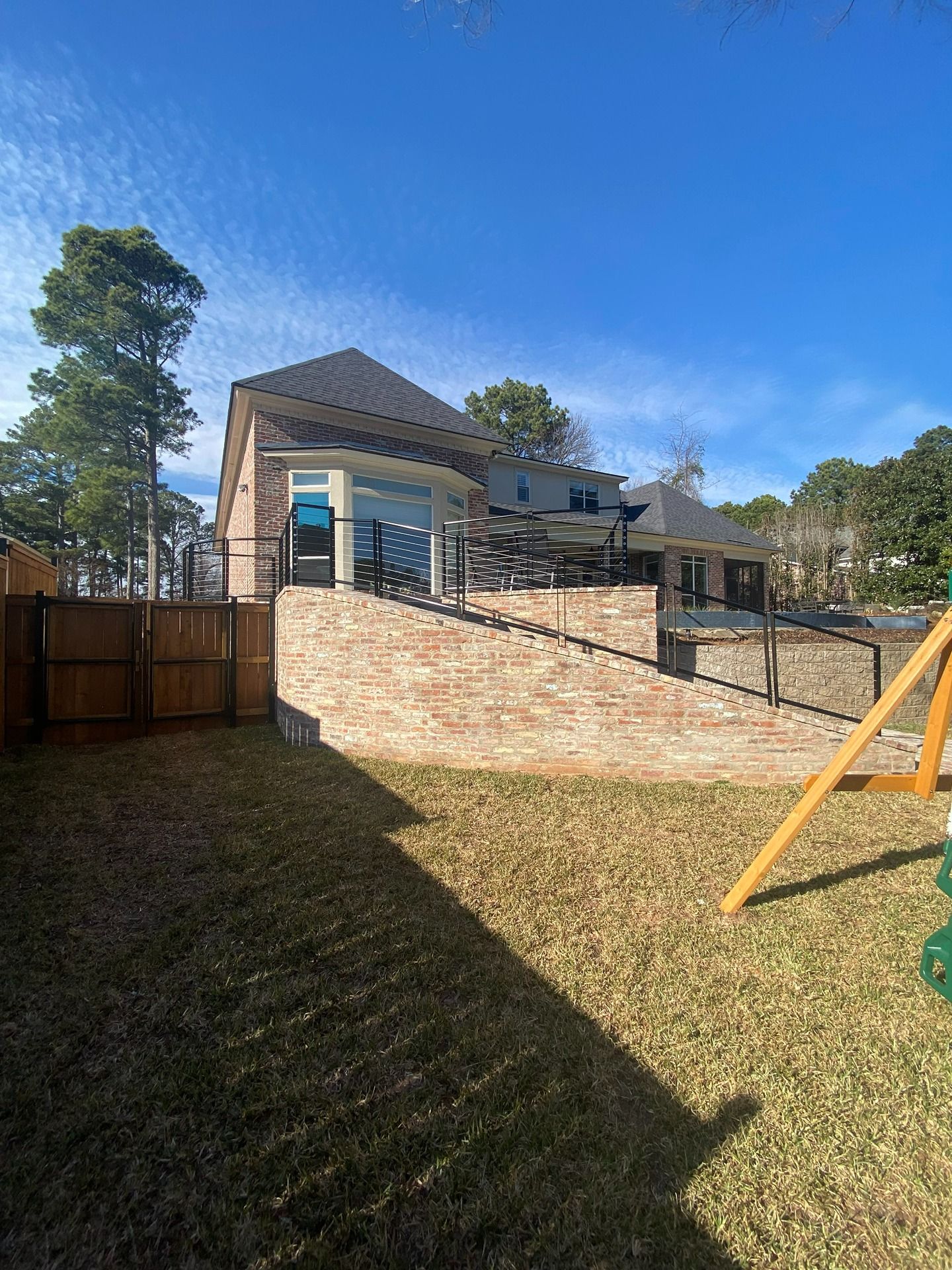 A large, two-story brick house with a raised stone terrace, metal railings, and a fenced yard under a clear blue sky.