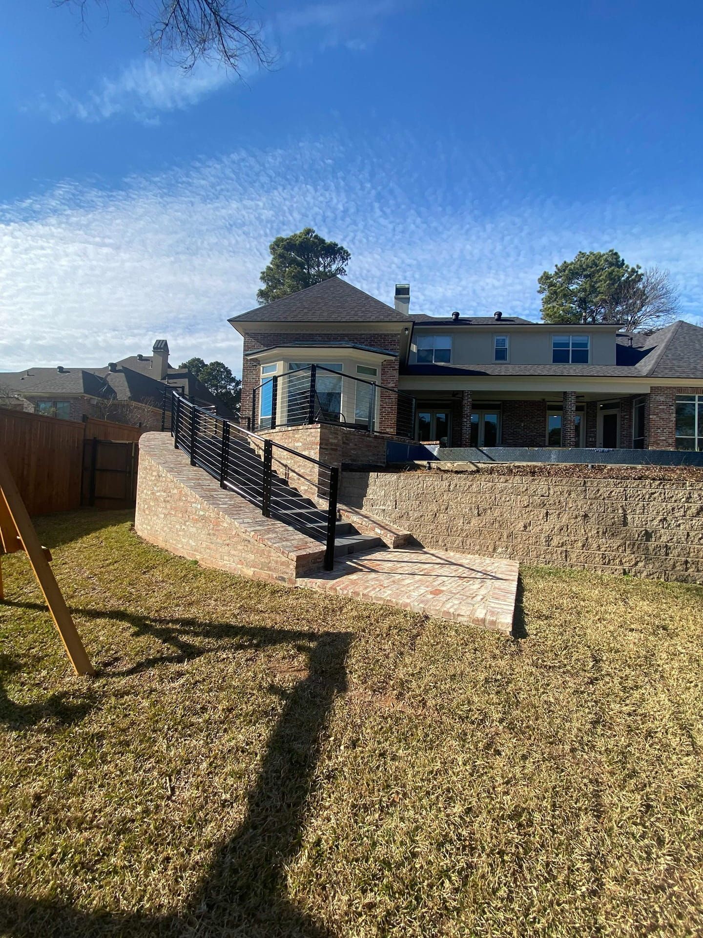 Outdoor stairs made of light stone with black railings leading up to a large brick house in a backyard setting.