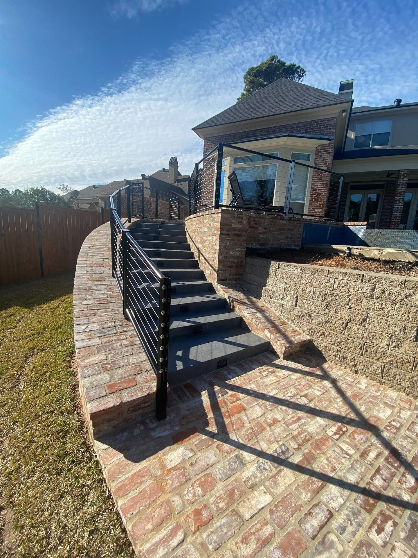 A stone staircase with black metal railings leads from a brick patio up to a house terrace on a sunny day.