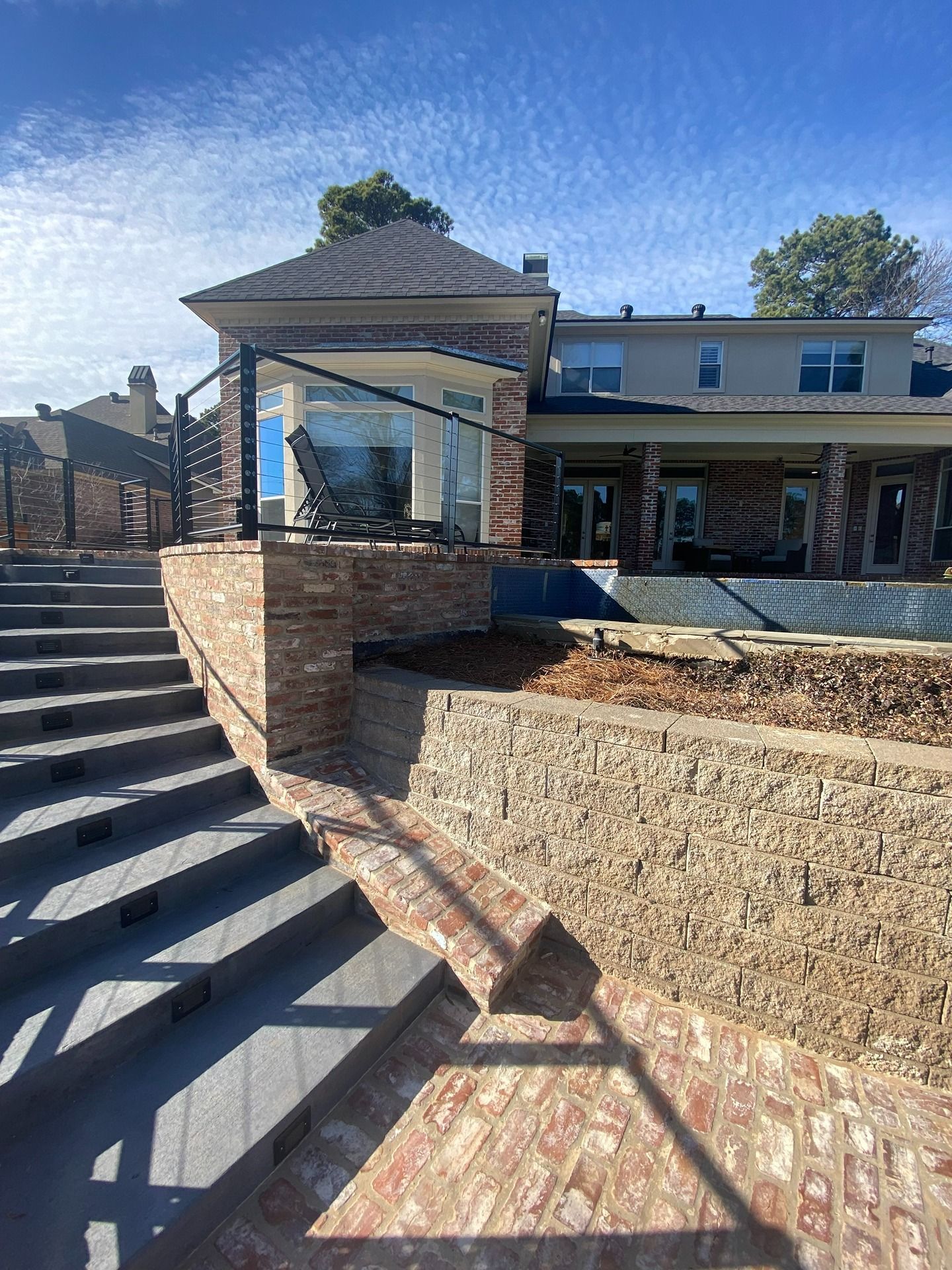 Outdoor stone steps lead to a house patio with a dark railing, stone retaining wall, and brick-paved lower area.