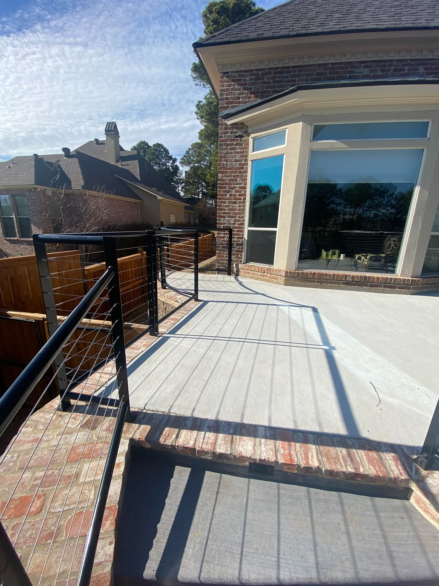 A raised patio featuring light gray deck boards, a black metal cable railing, and a brick border next to a brick home.