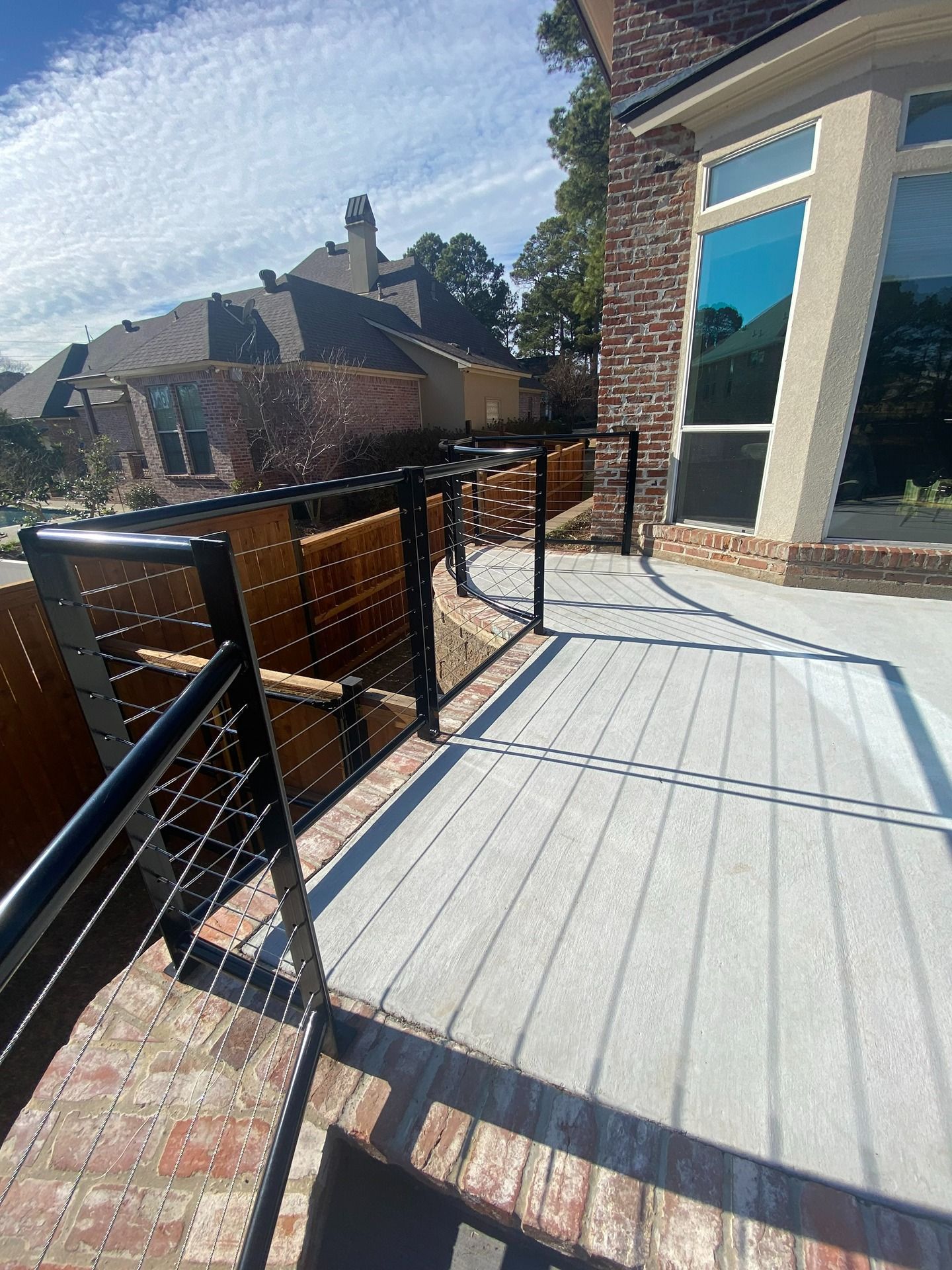 A concrete patio with a black metal cable railing overlooking a residential backyard under a sunny blue sky.