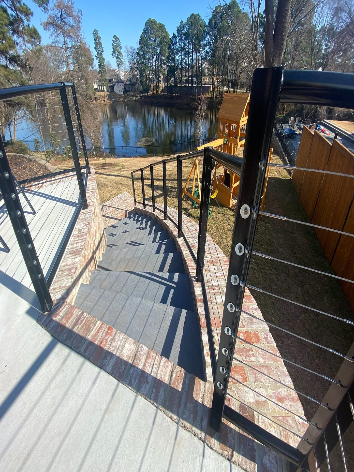 Curved stairs with grey decking and black metal cable railings lead down toward a grassy yard and a pond.