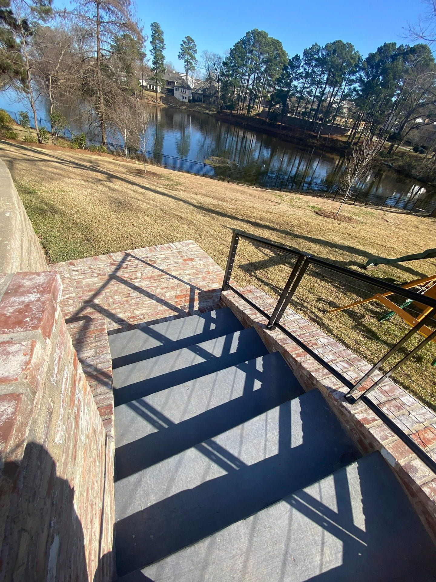 A high-angle view of outdoor stone stairs leading down to a lawn and a calm lake on a sunny day.