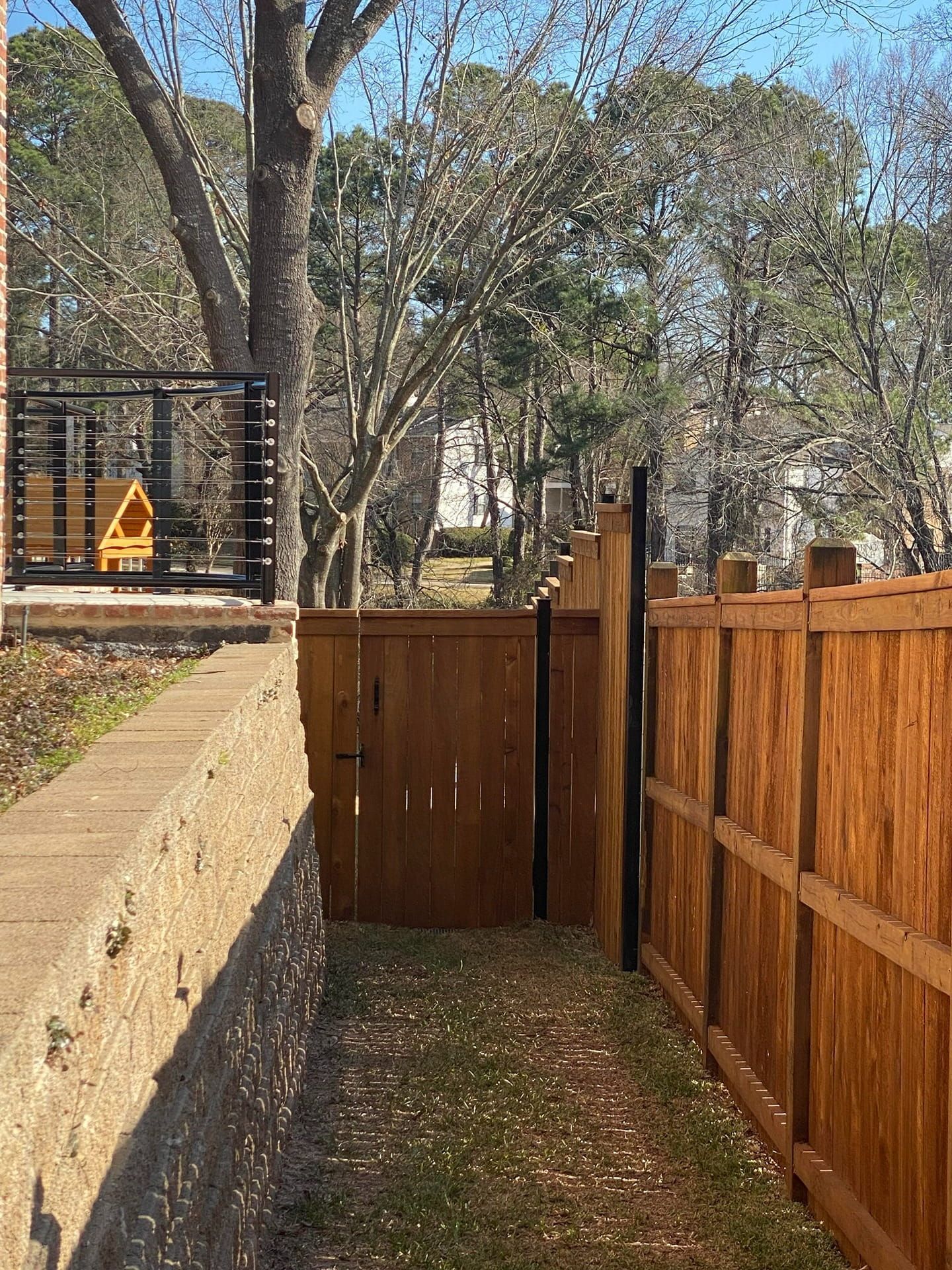 A side yard with a tan brick retaining wall on the left and a wooden fence with a gate on the right, leading to trees.