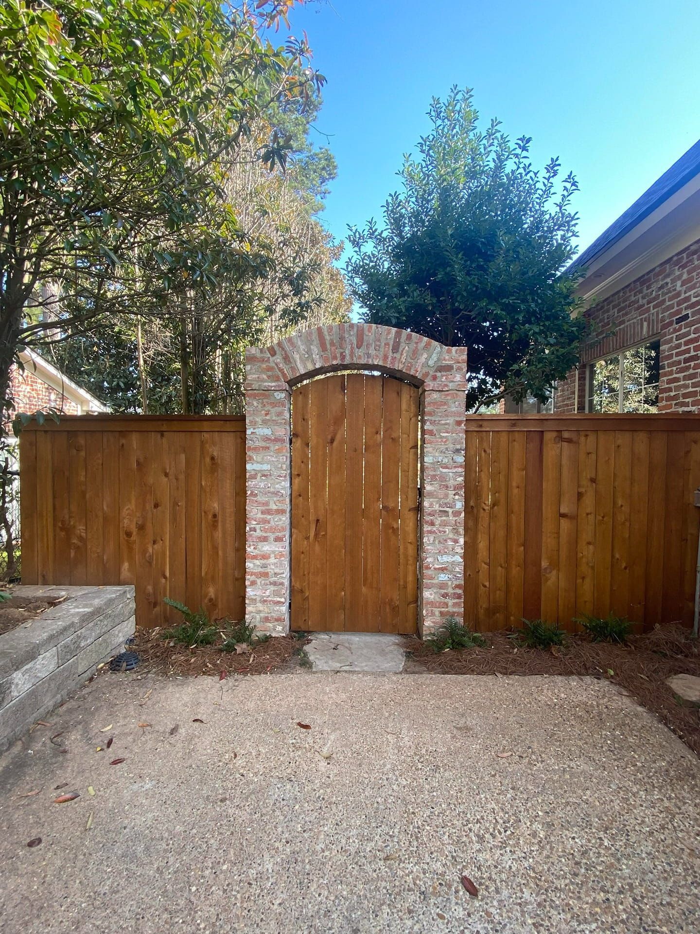 A wooden gate set into a brick archway between two wooden fences, viewed from a pebble patio on a sunny day.