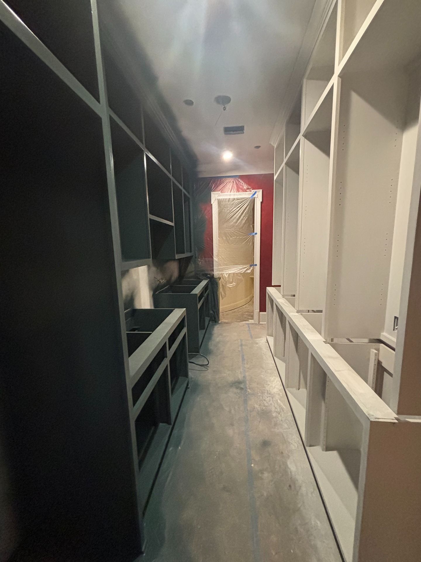 A narrow hallway under construction featuring dark cabinets on the left, white units on the right, and a red door ahead.