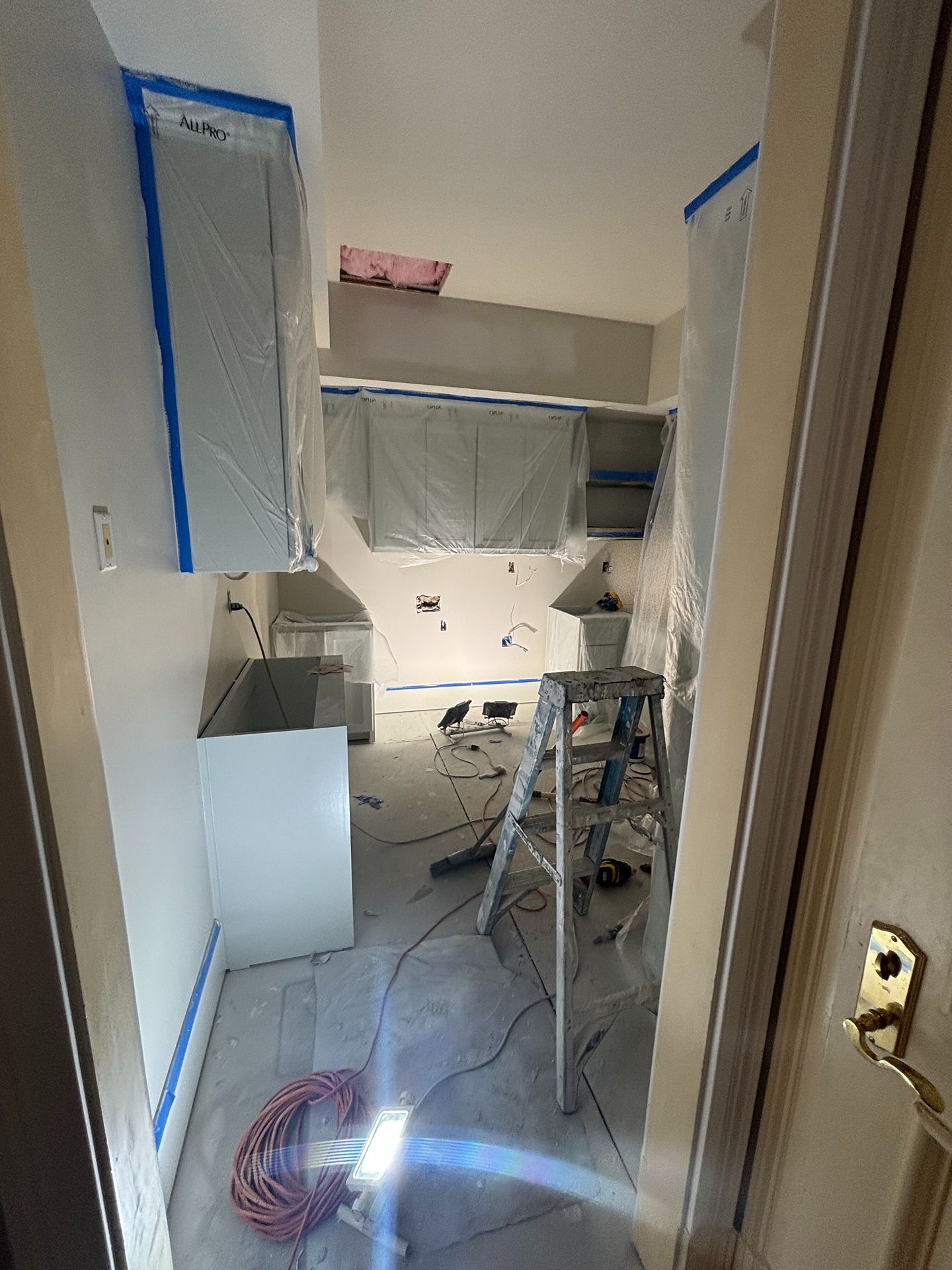 A small kitchen undergoing renovation, featuring cabinets covered in plastic, a ladder, and tools on the floor.