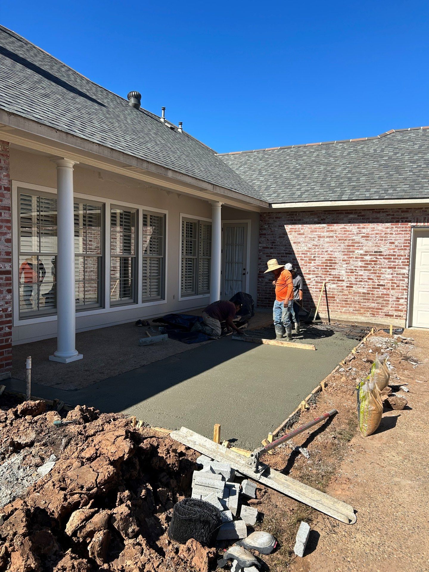 Workers smooth freshly poured concrete for a patio outside a brick house with columns.