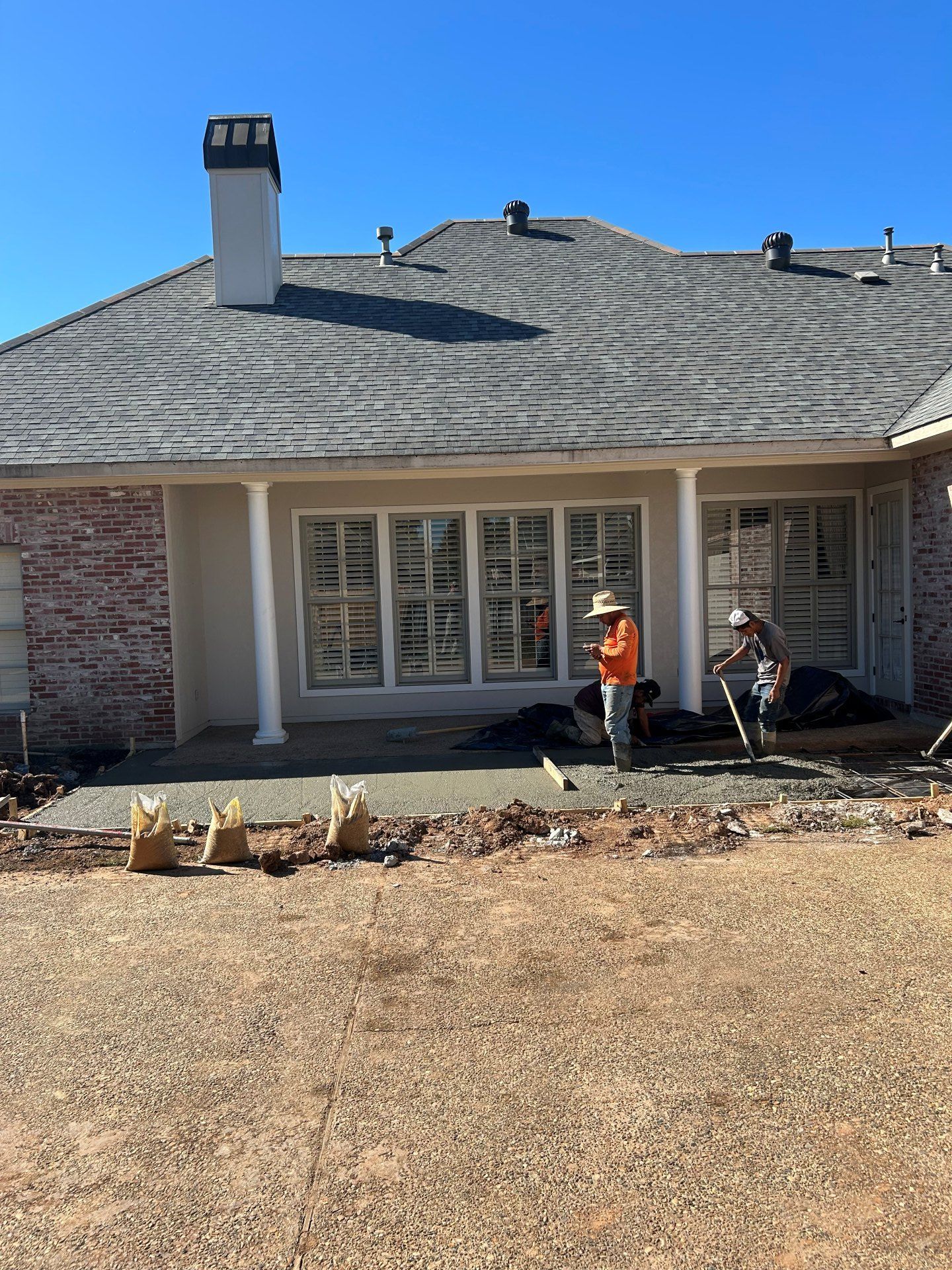 Two workers in high-visibility vests work on a new concrete patio in front of a brick house with a grey shingled roof.