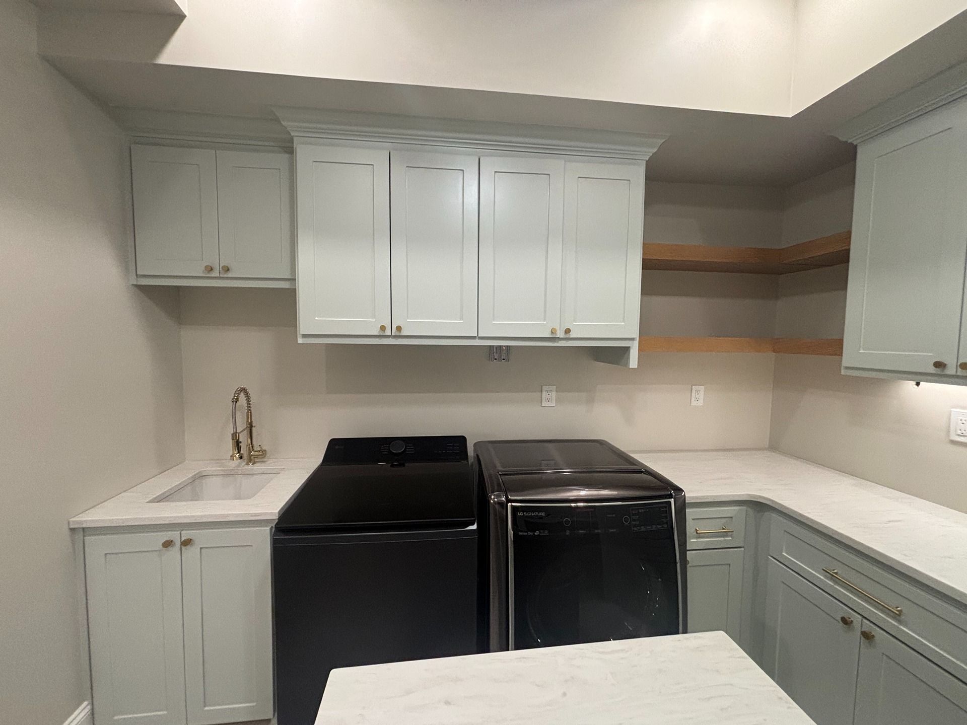 A laundry room with light blue cabinets, dark appliances, a white countertop with a sink, and open wood shelving.