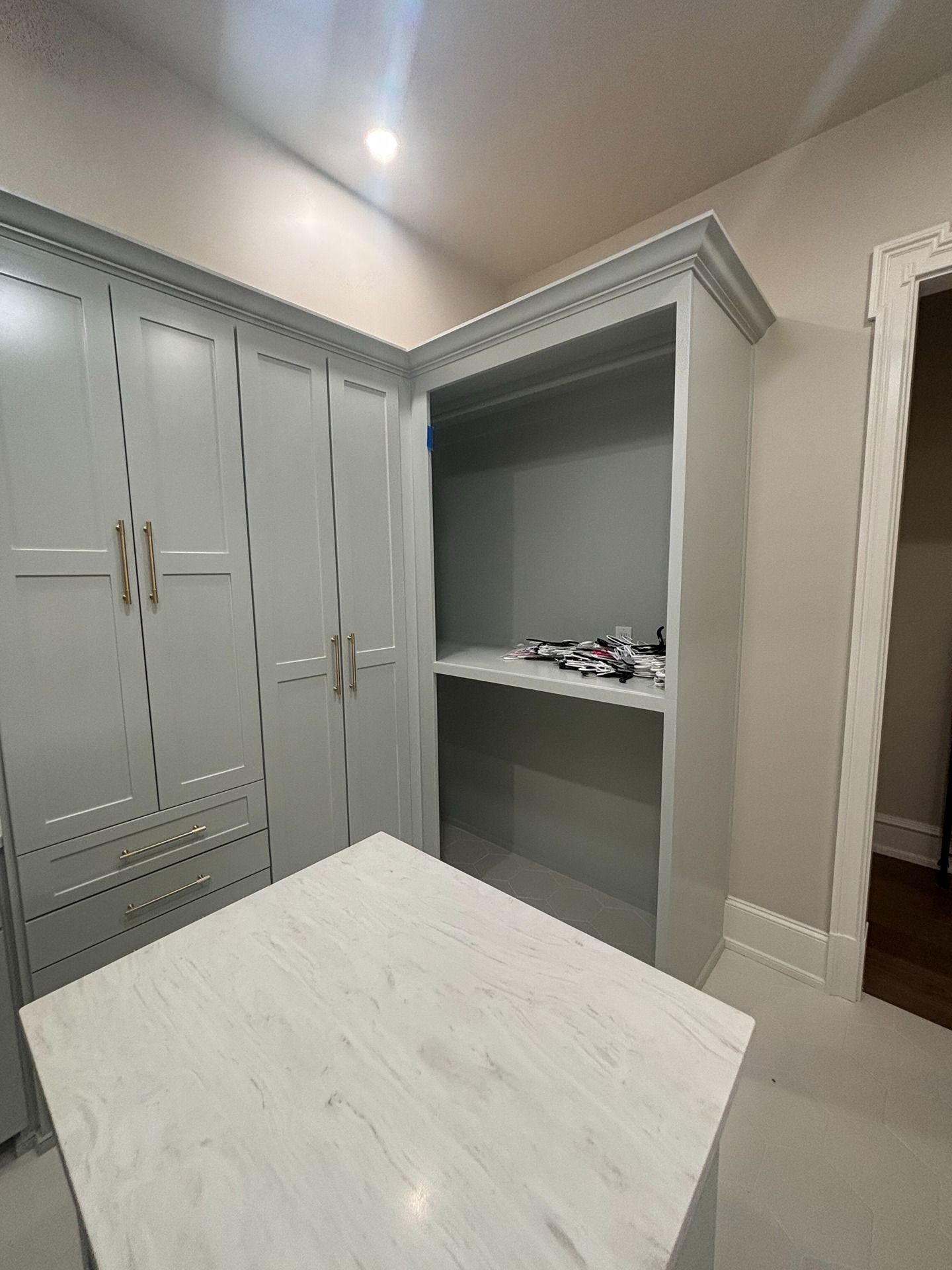 A walk-in closet featuring light gray cabinetry, a white marble-topped island, and an open shelving unit with shoes.