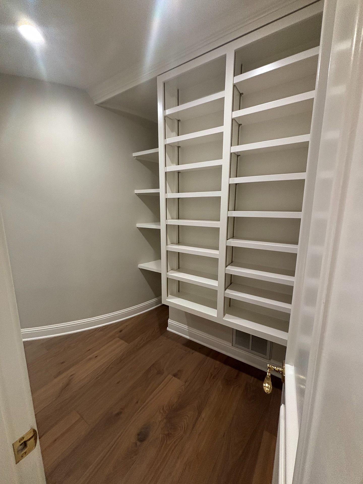 A white built-in shelving unit with multiple shelves sits in a walk-in closet with dark wood floors and grey walls.