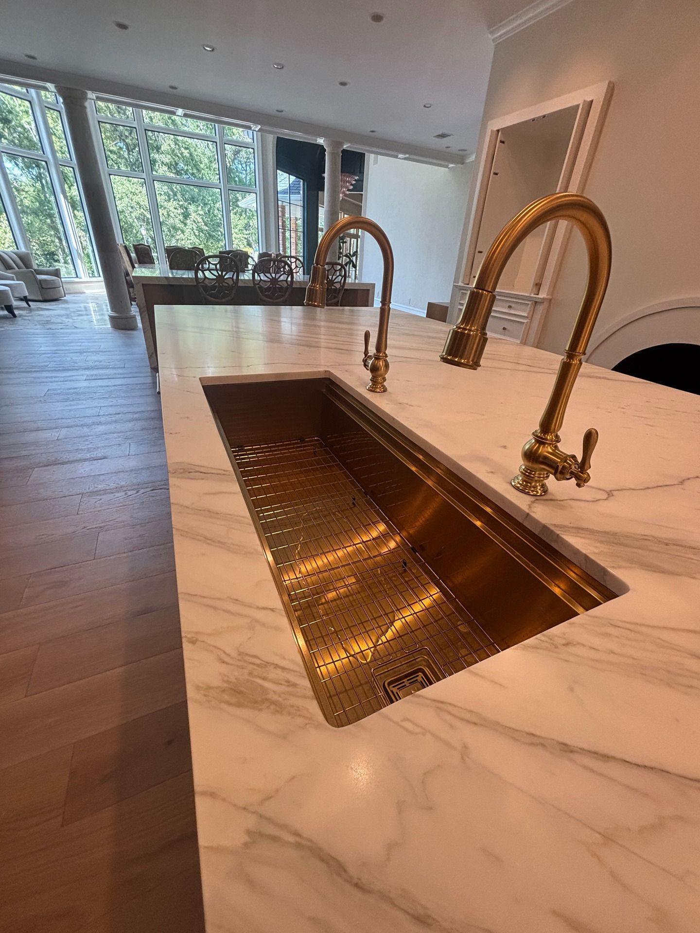 A brass kitchen sink with two matching gold faucets set into a white marble kitchen island in a bright, modern room.