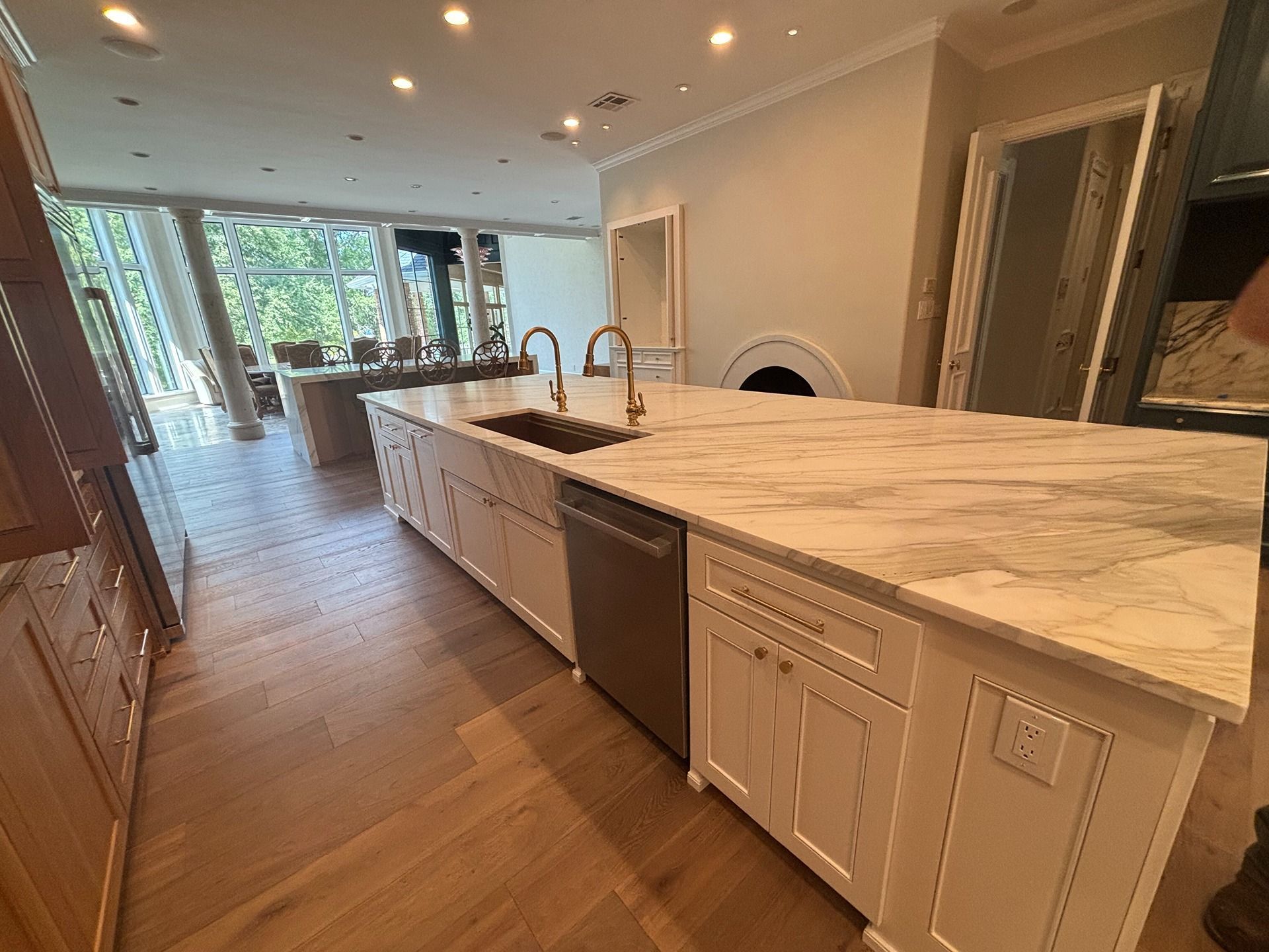 A modern, bright kitchen featuring a long white marble island with a sink, stainless dishwasher, and wood flooring.