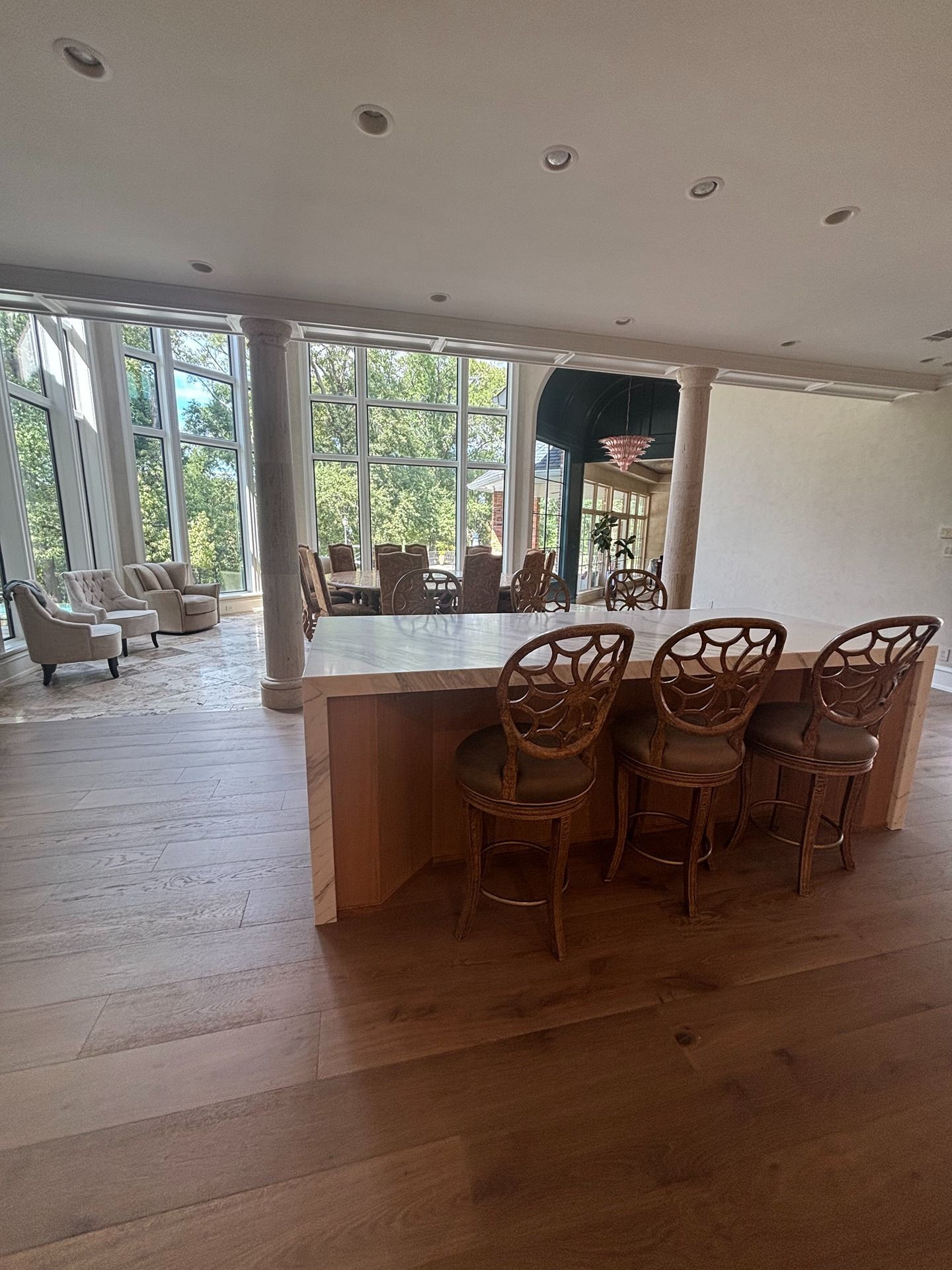 Modern kitchen island with three ornate metal stools on light wood floors, opening to a sunlit dining area with views.