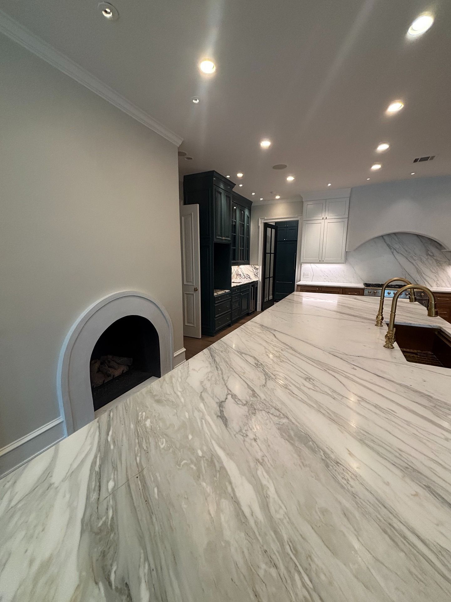 A kitchen island with white marble countertops, a curved fireplace, and dark blue cabinets under recessed lighting.