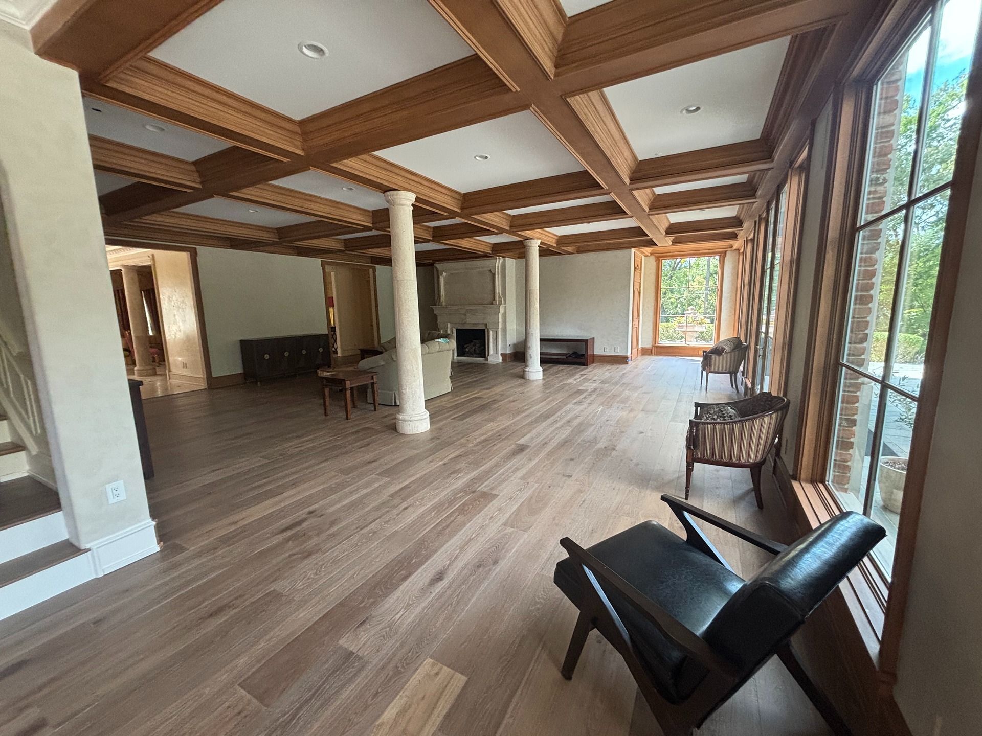A spacious room with light hardwood floors, a coffered wood ceiling, two white pillars, and leather and fabric chairs.