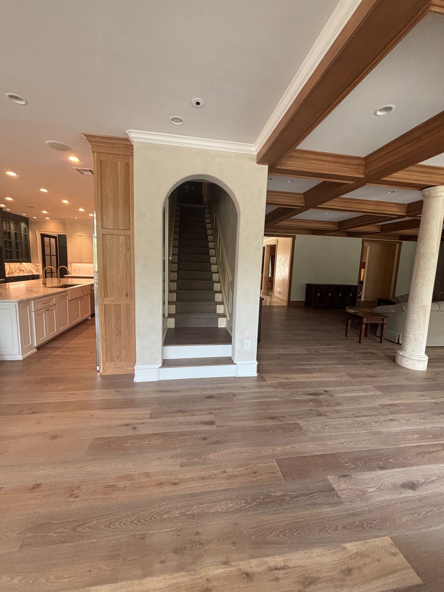 A spacious home interior with light wood floors, an arched staircase entry, a kitchen on the left, and ceiling beams.