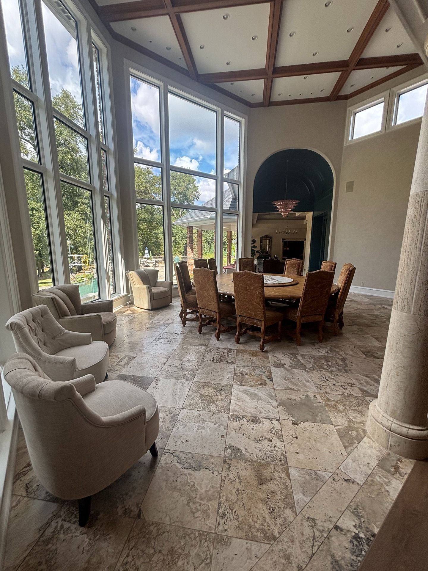 A high-ceilinged room with large windows, stone floors, a dining table with chairs, and accent armchairs.