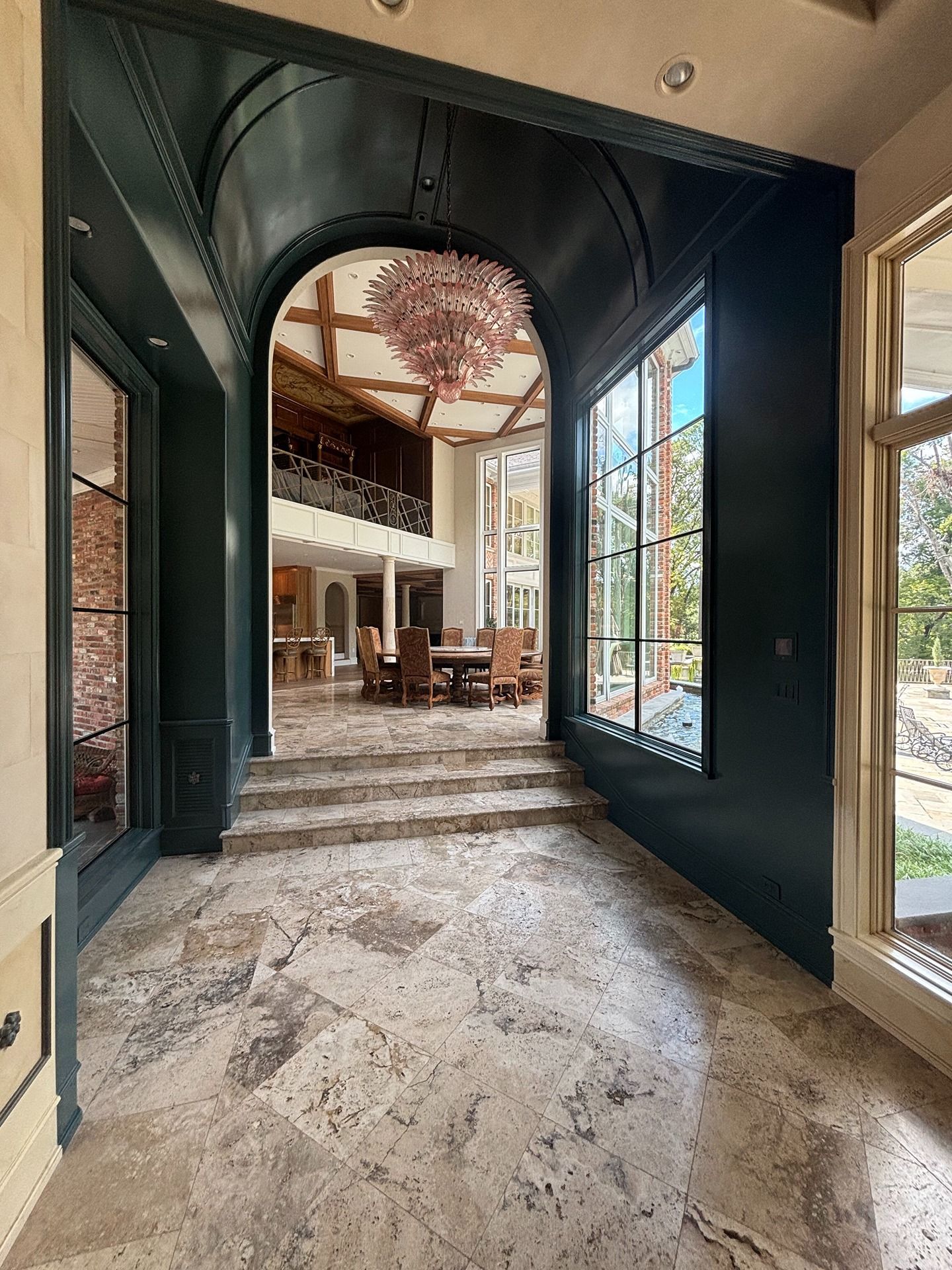 A tiled entry hall with a dark arched ceiling leads through steps toward a spacious room with wooden beams and a chandelier.