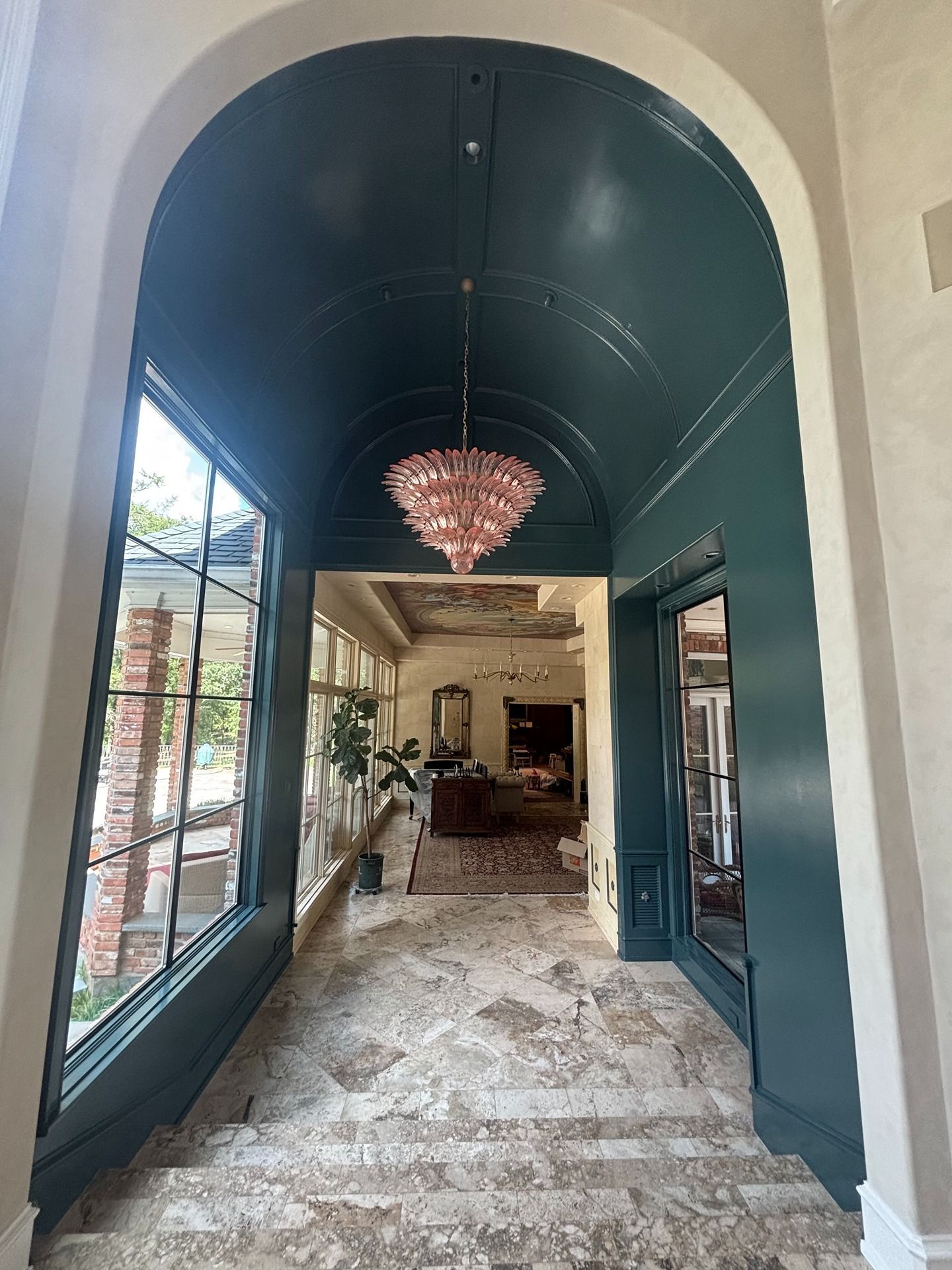 A teal arched hallway with a chandelier, large windows overlooking a patio, and stone flooring leading into a home.