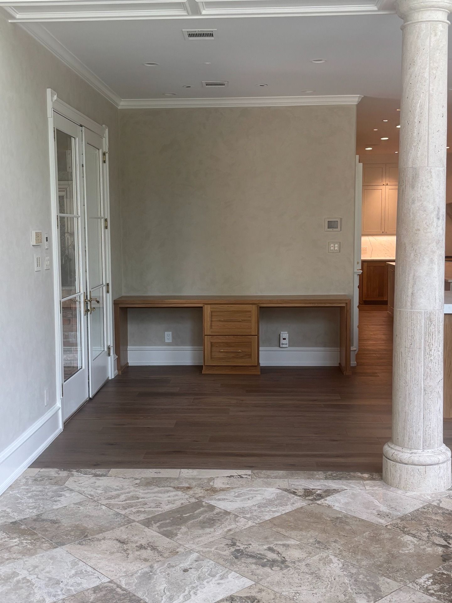 A wooden desk with drawers sits against a beige wall in a hallway with a stone pillar and marble flooring.