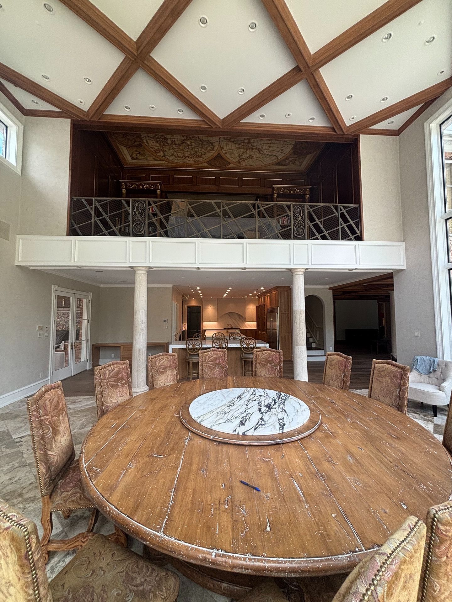 A round wooden table with a marble lazy susan sits in a room with a high ceiling, exposed beams, and an indoor balcony.
